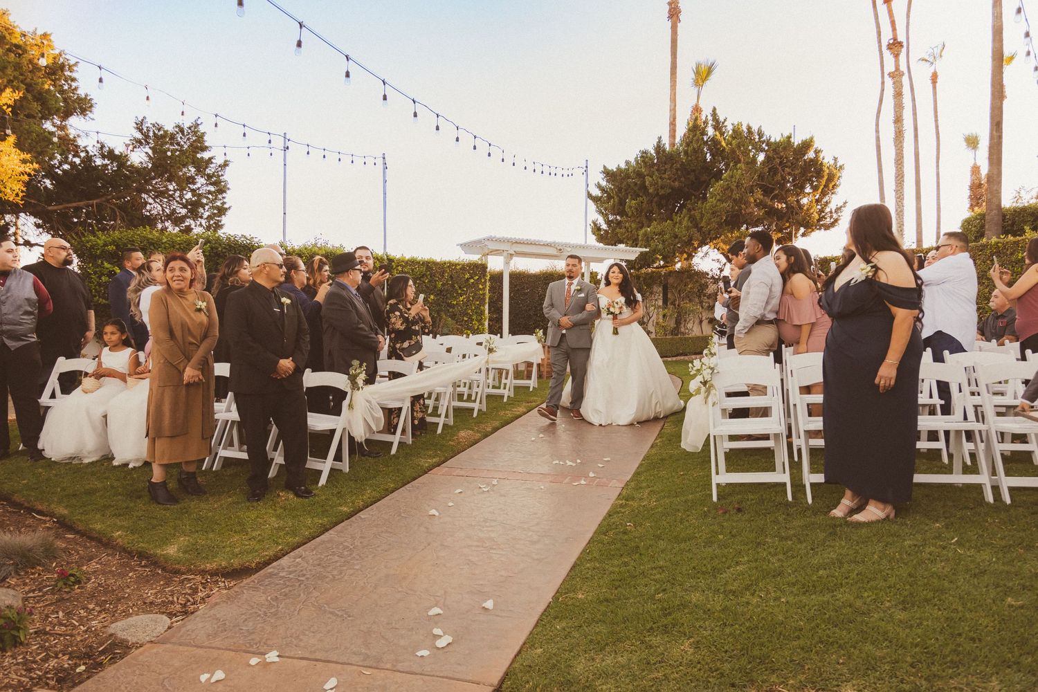 Garden wedding ceremony with guests seated on white chairs beneath string lights at dusk.