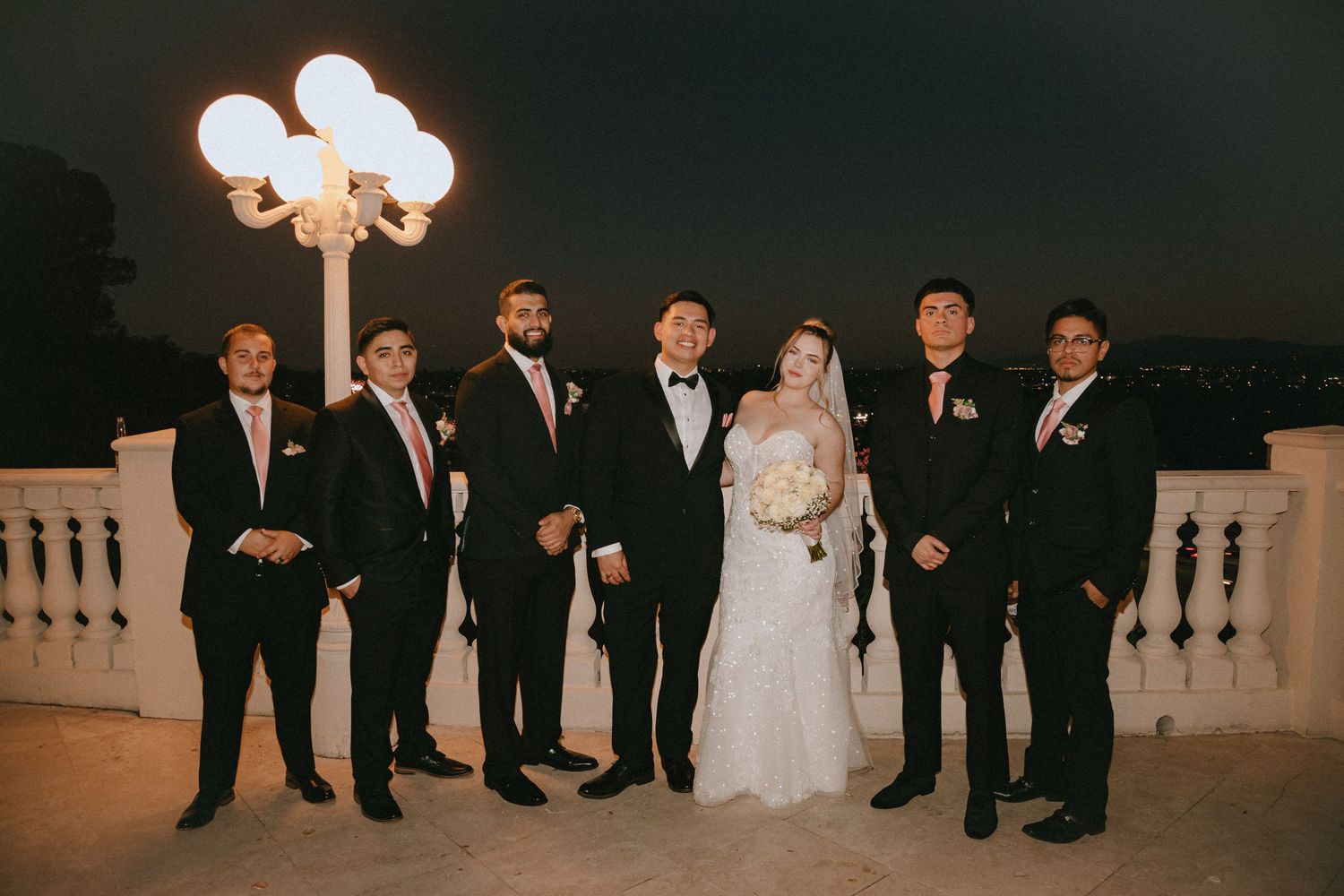 Wedding party group photo with elegant lighting and balustrade backdrop.