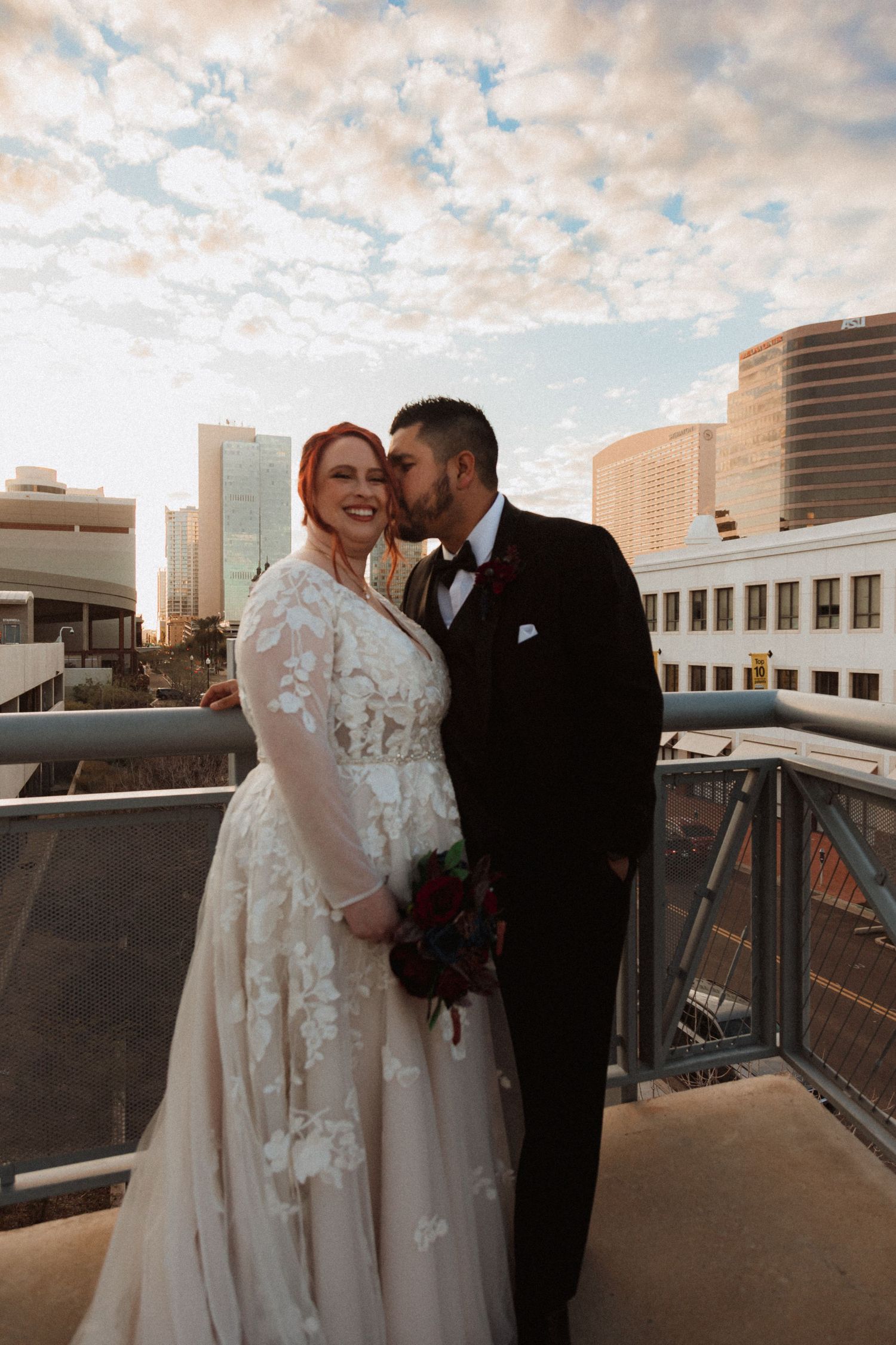 Wedding couple shares joyful moment on urban rooftop with dramatic sunset sky.