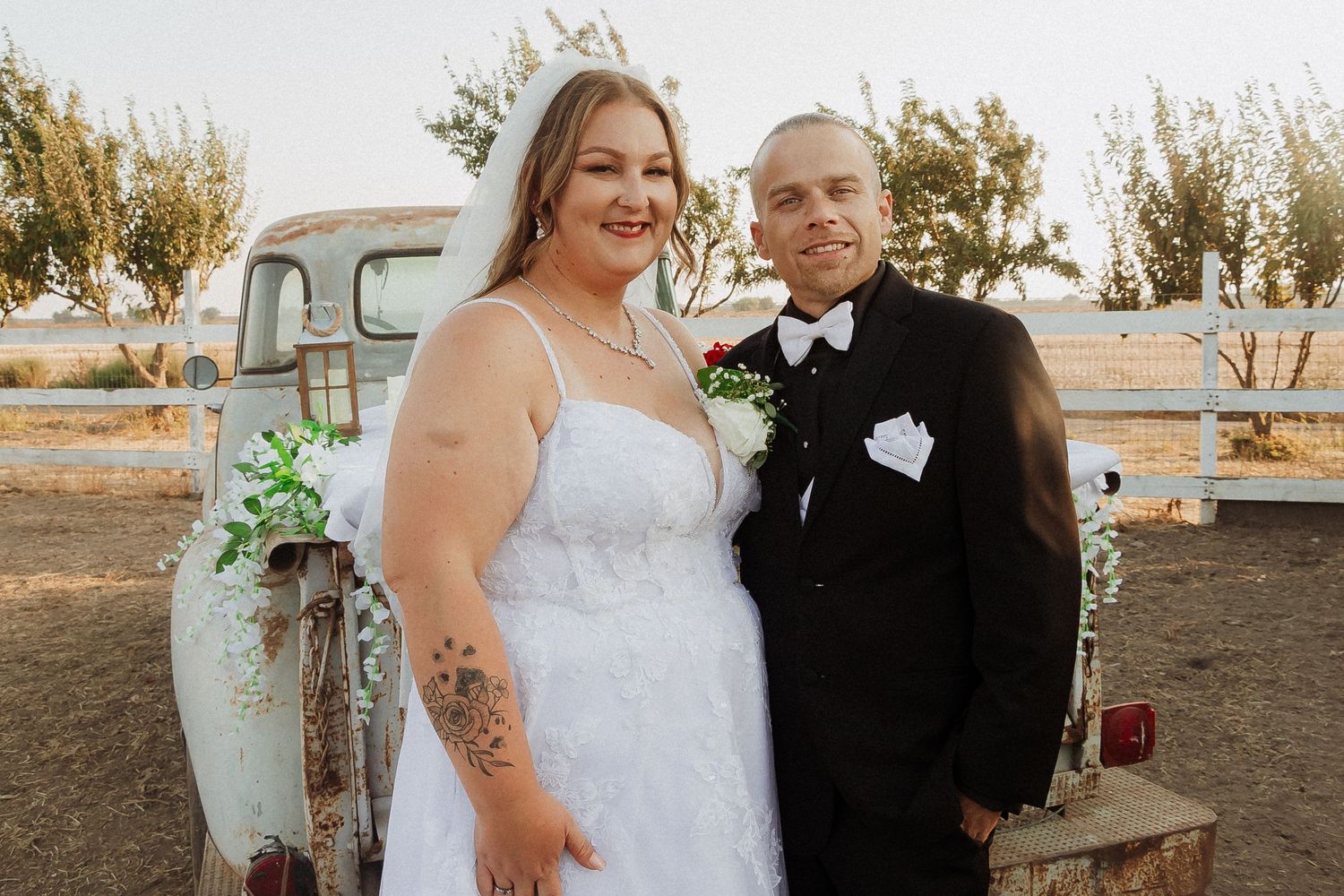 A happy couple poses together in formal wedding attire with a classic light blue pickup truck in the background.