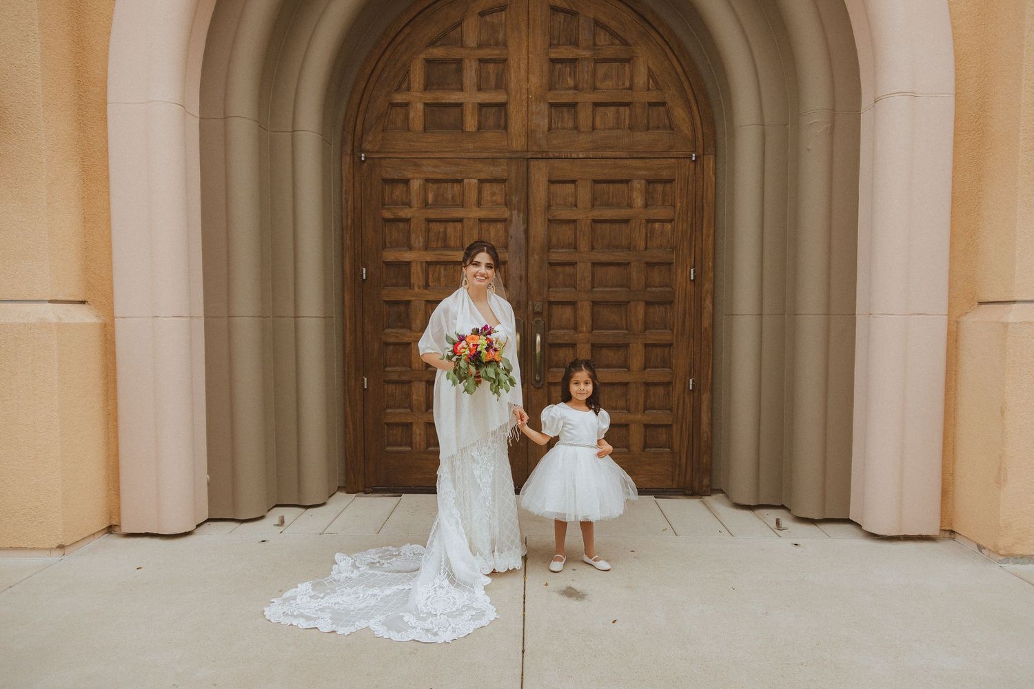 A bride in a white lace gown stands with a young flower girl in front of an arched wooden church door.