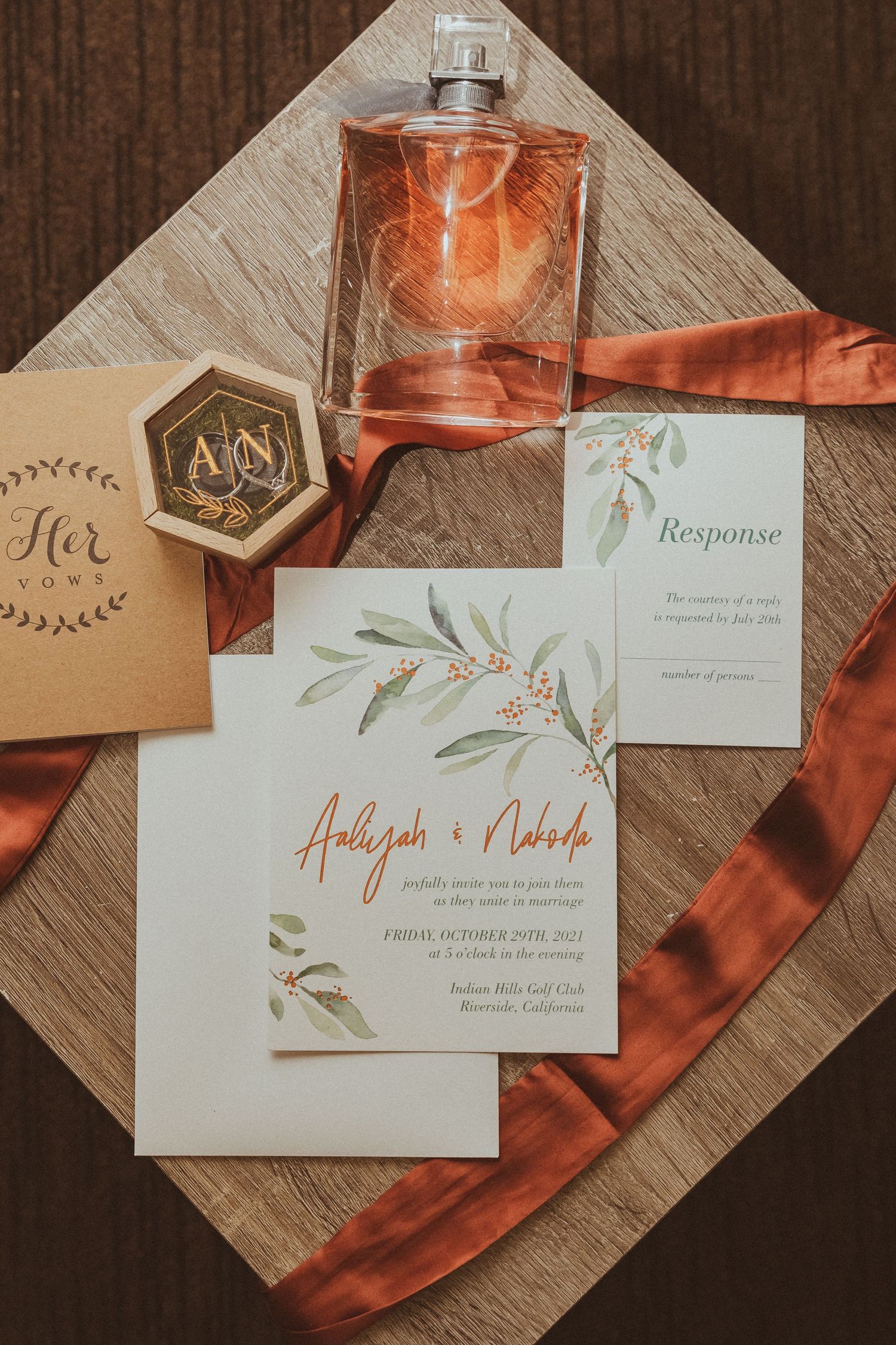 Elegant wedding invitation with olive branch design, perfume bottle and terracotta ribbon on wooden surface.