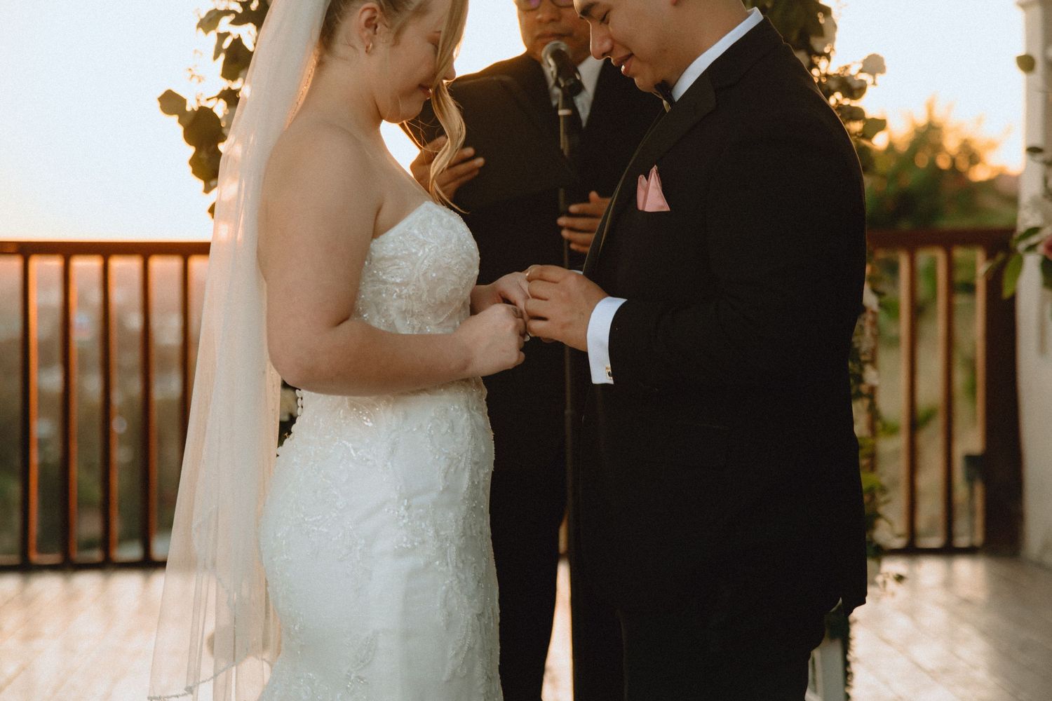 A couple exchanges rings during their wedding ceremony under a floral arch at sunset.