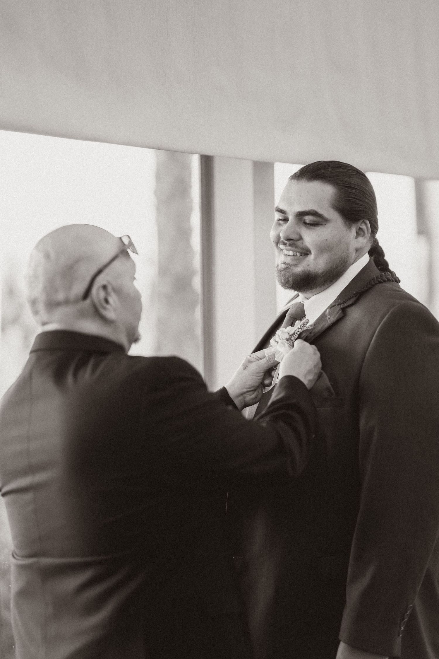 A black and white photo of someone helping adjust another person's boutonniere before a formal event.