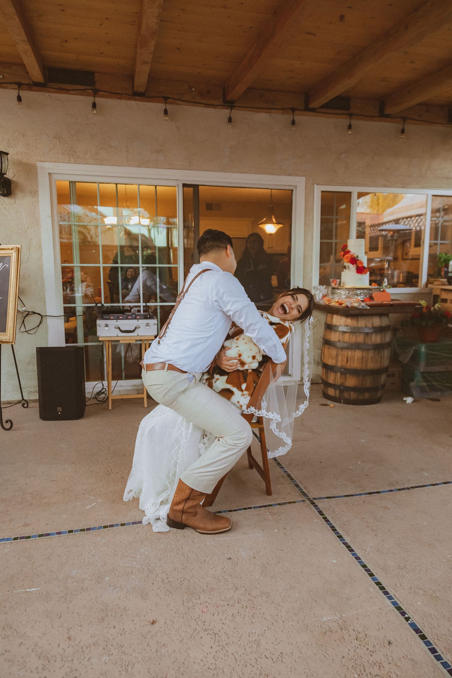 A couple dances playfully during a wedding reception on an outdoor patio with wooden beams overhead.