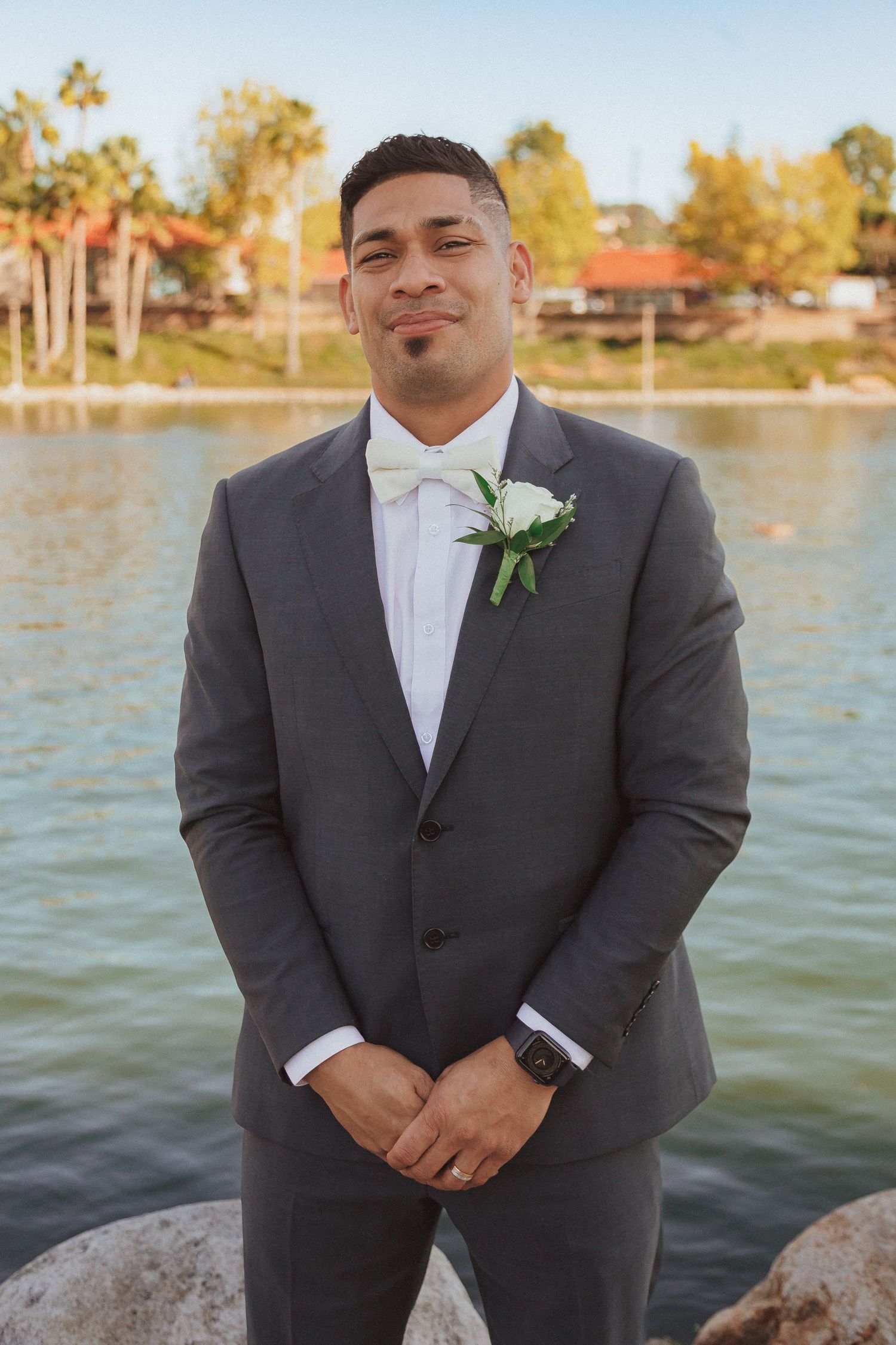 Groom in formal grey suit and white bowtie poses by lakeside.