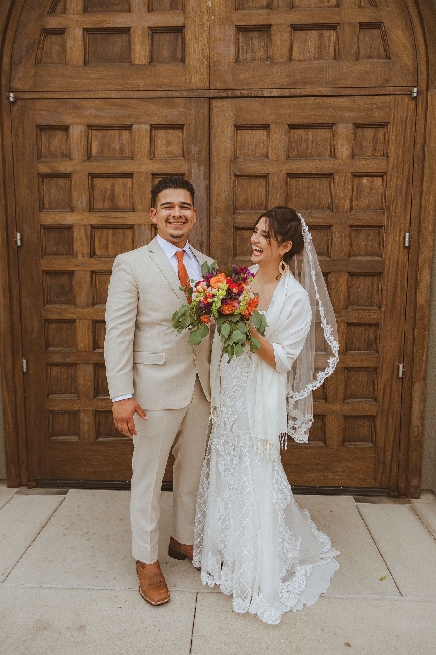 A bride and groom pose together in front of a wooden door, with the bride holding a vibrant orange and red bouquet.