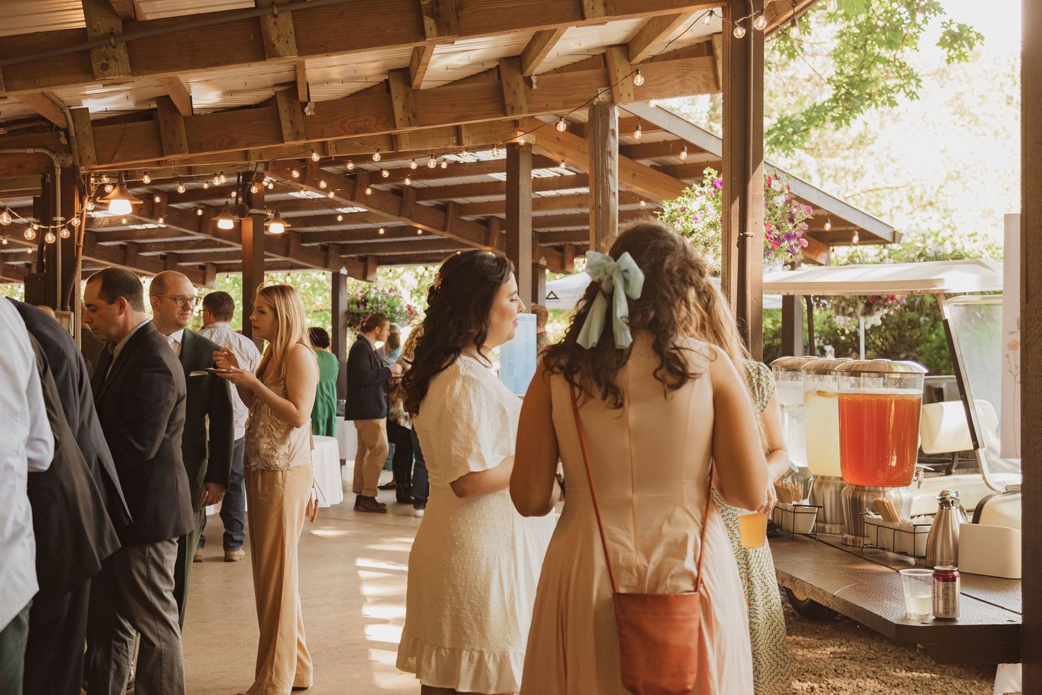 Wedding guests gather under a wooden pergola during a daytime outdoor reception.