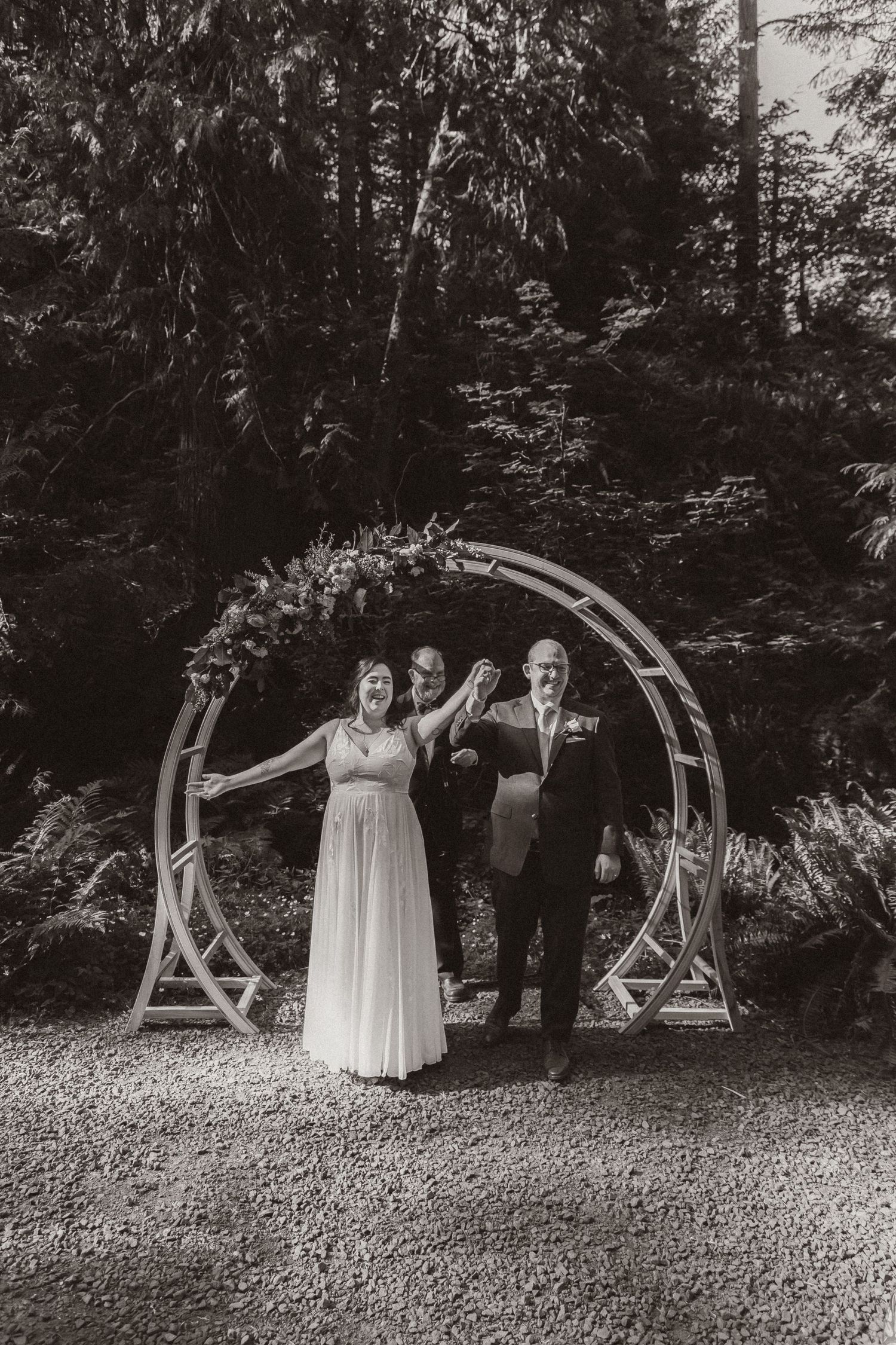 Black and white capture of celebratory moment under floral arch during forest ceremony.