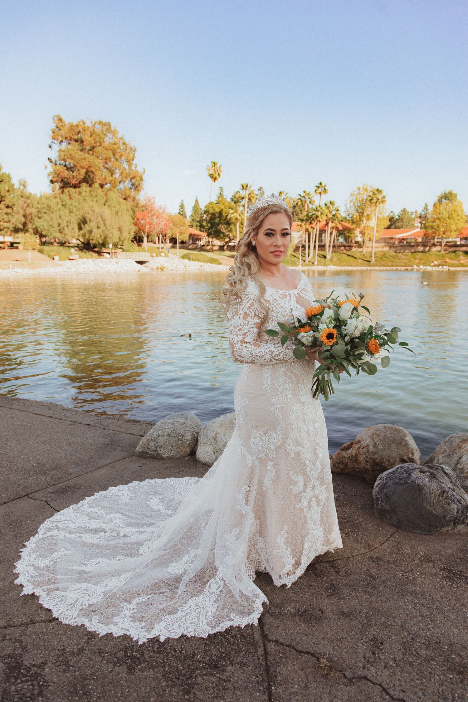 Bride in long-sleeve lace gown poses with fall bouquet near lakeside rocks.