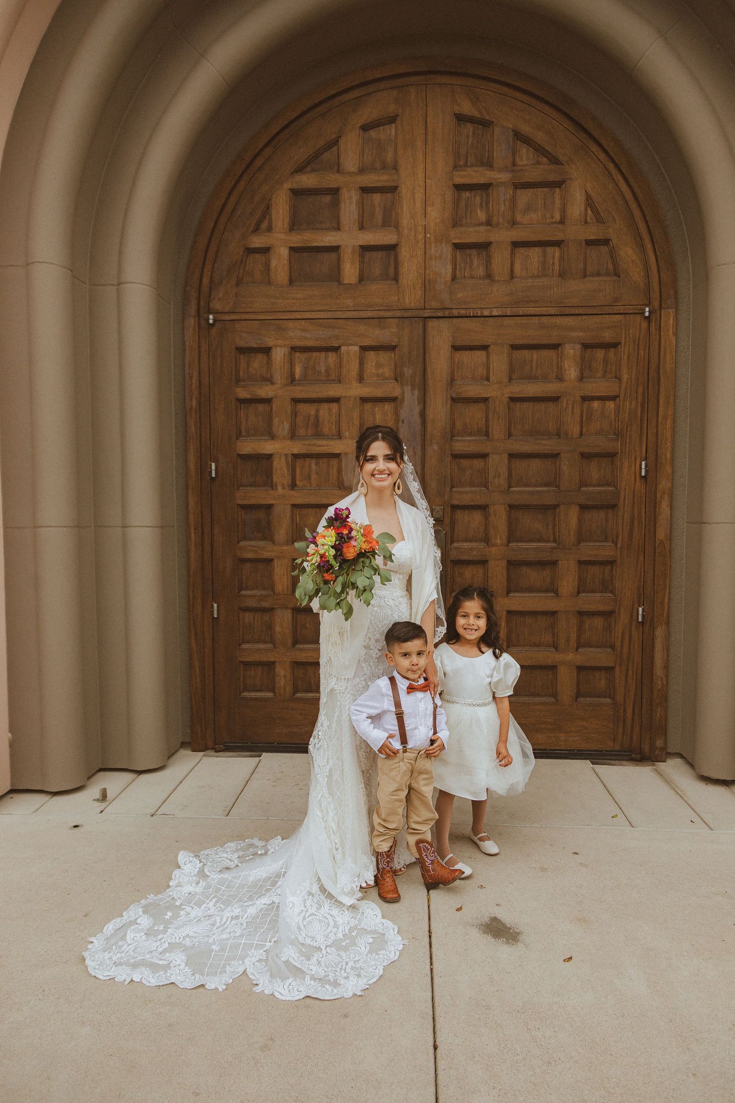 Wedding portrait in front of ornate wooden church doors featuring a bride and young attendants.
