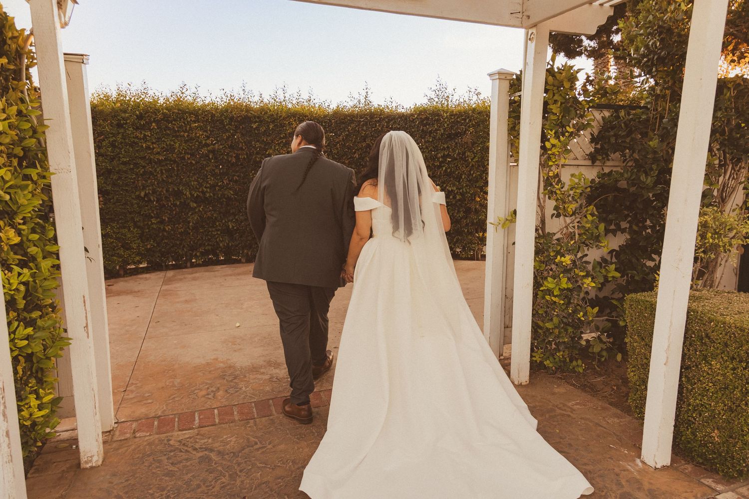 A wedding couple walks through a garden archway during sunset.