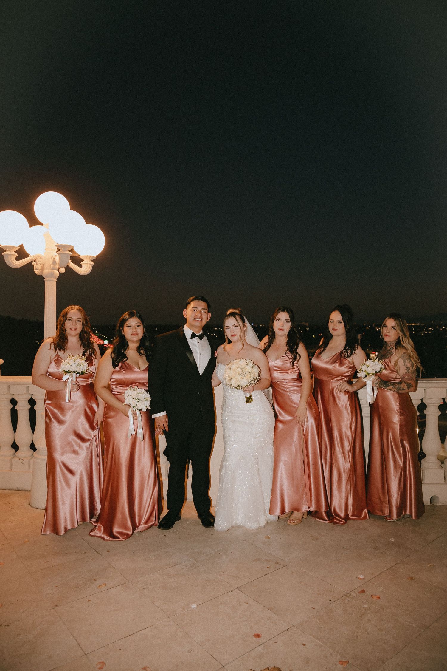 Wedding group photo featuring bridesmaids in shimmering rose gold gowns on evening terrace.