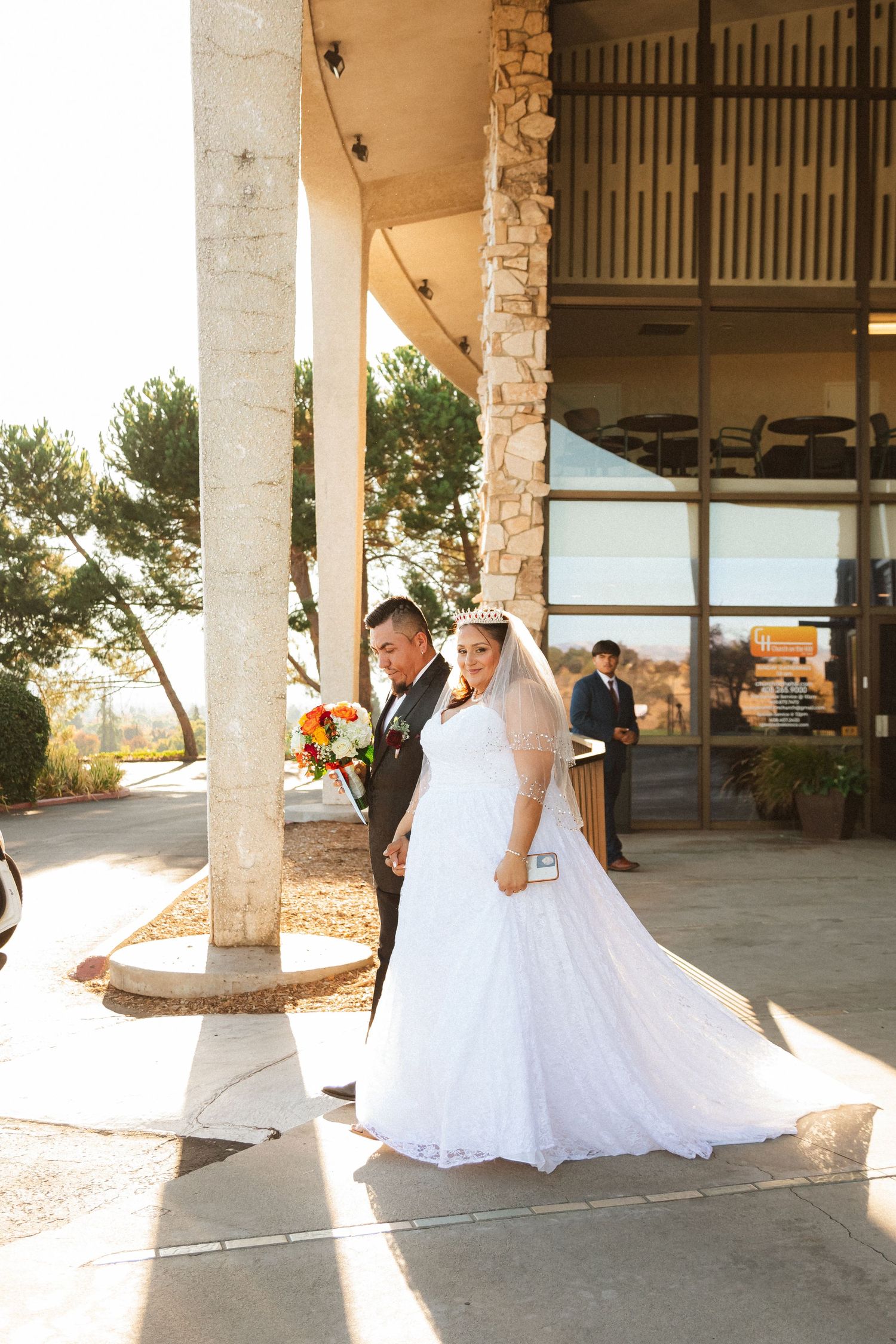 Newlyweds pose in golden sunlight outside a modern building with stone columns.