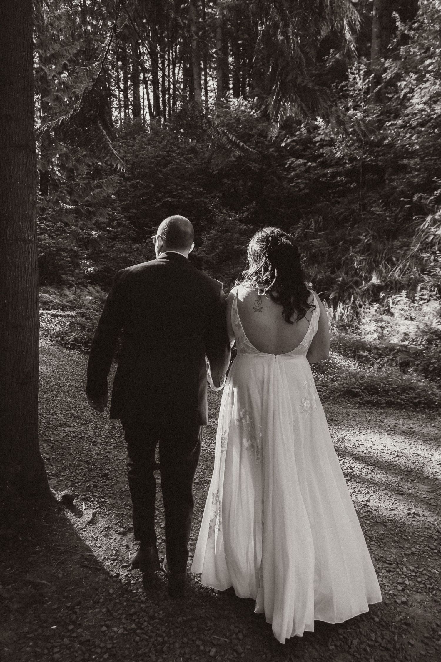 Black and white photo of newlyweds walking hand in hand through a wooded forest path.