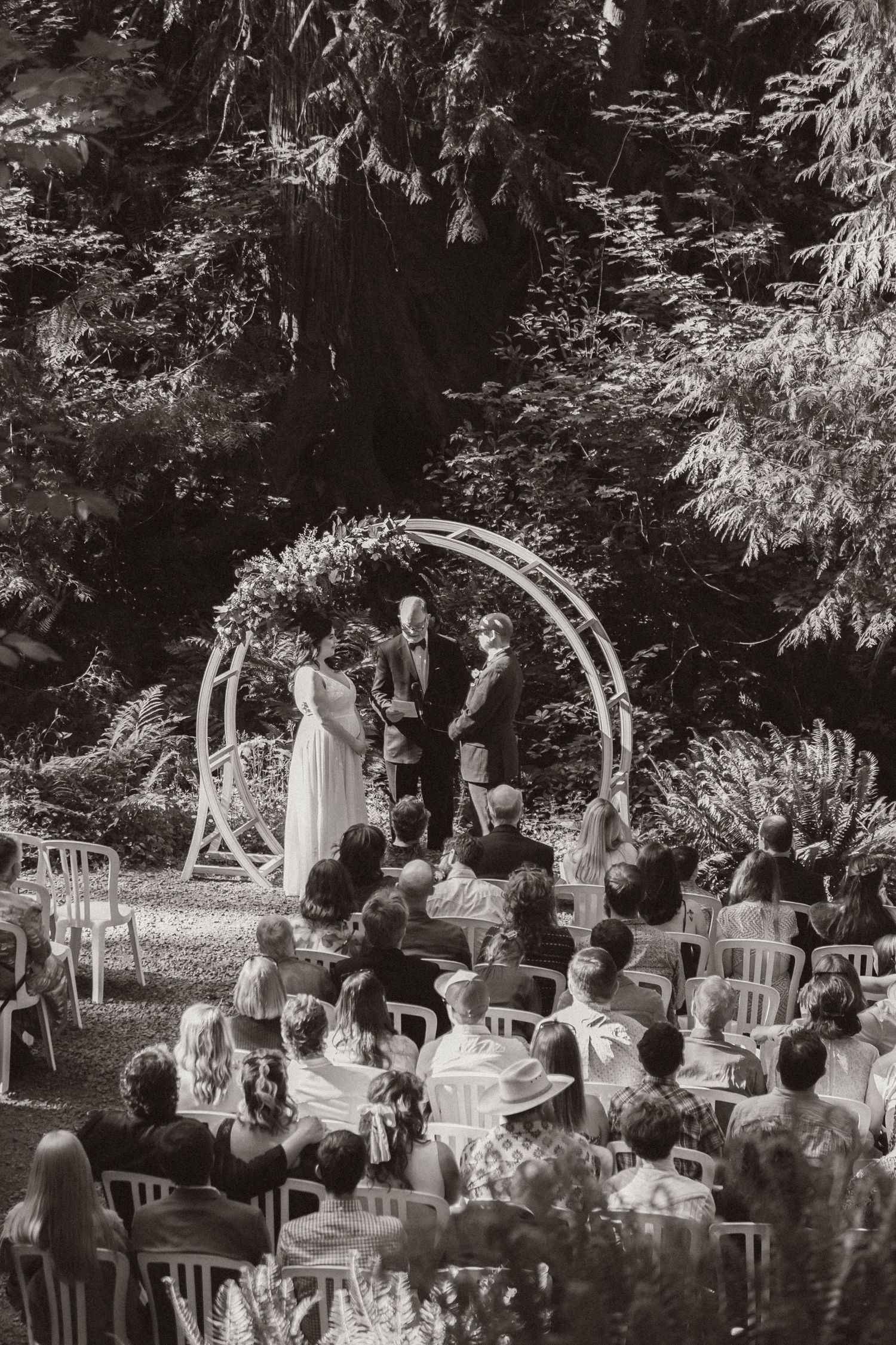 A wedding ceremony under an arch in a forest amphitheater with guests seated on white chairs.