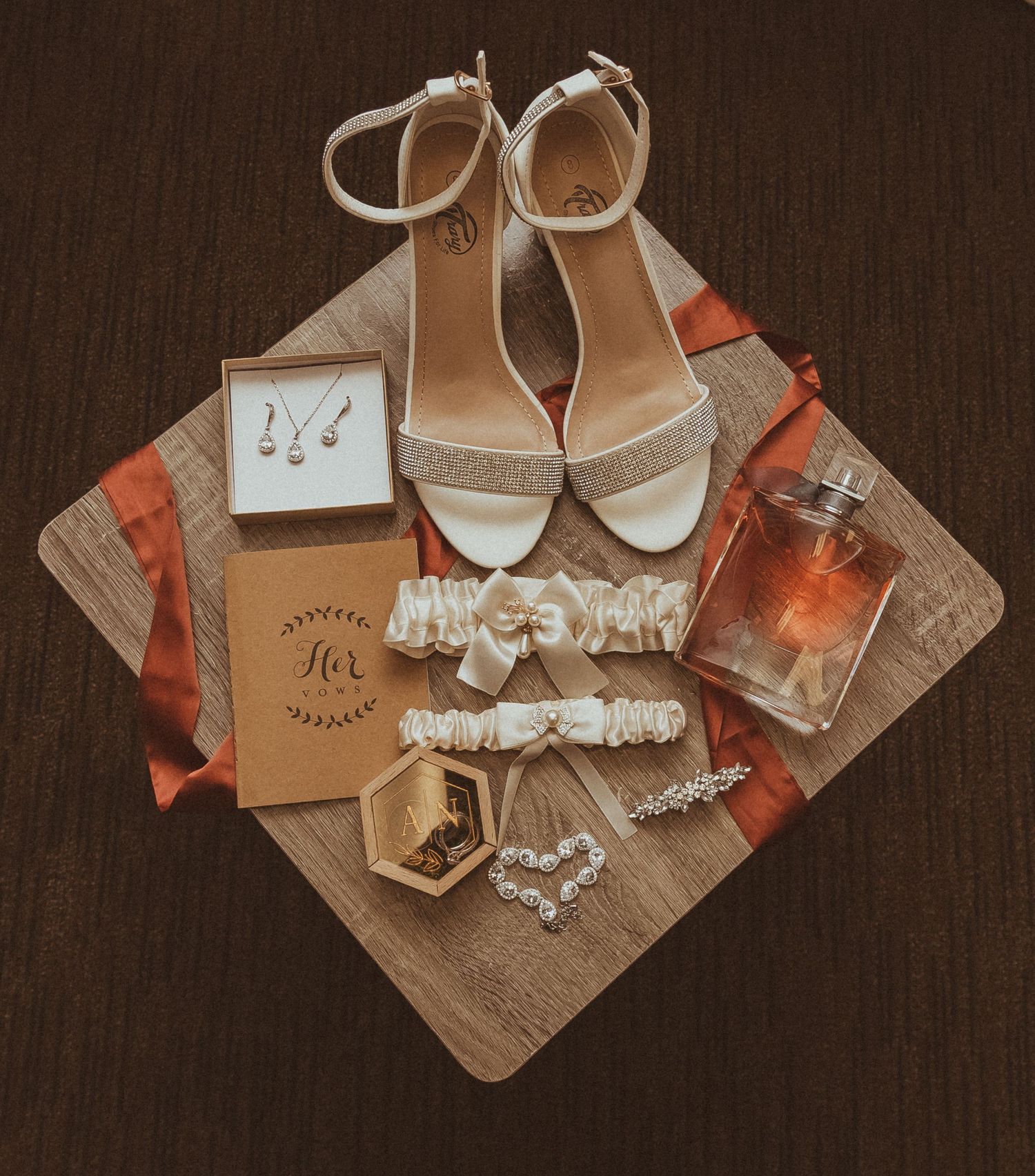 Wedding accessories including white sandals, garters, jewelry, and perfume arranged on wooden surface with orange accents.