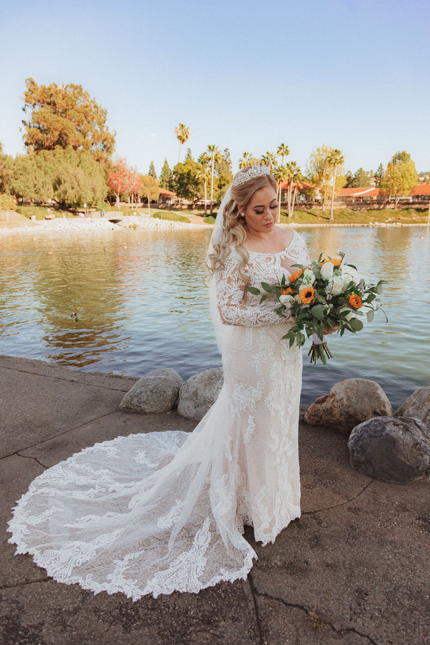 Full length view of bride in fitted lace gown with long train standing by scenic lake.