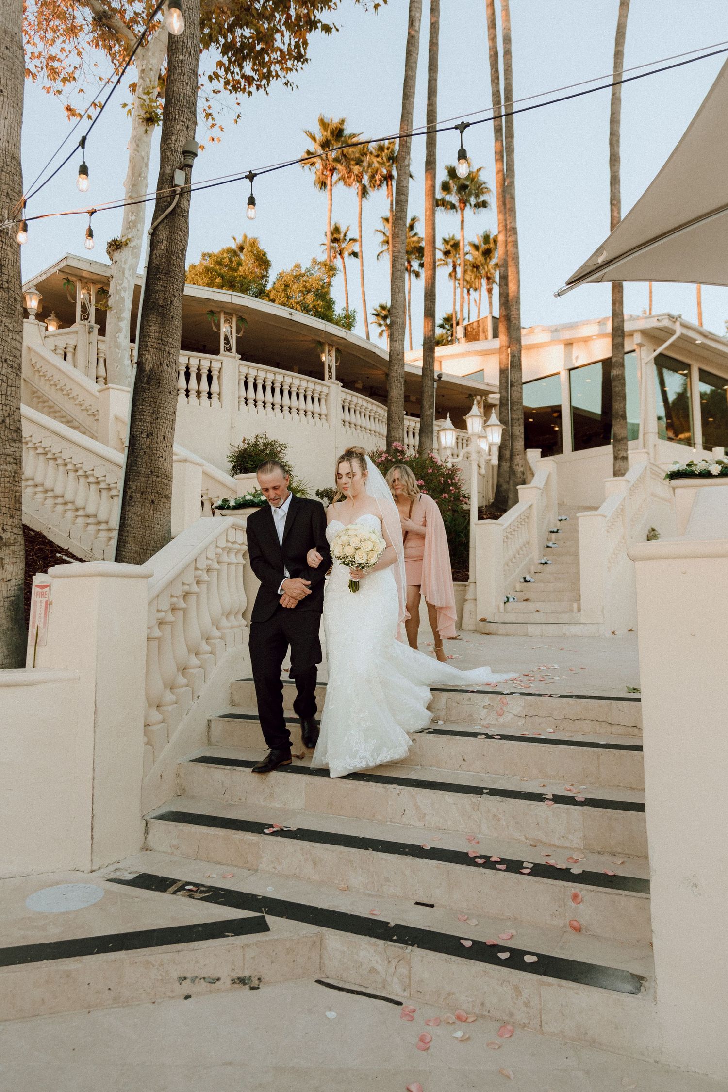 A formal wedding party descending elegant white concrete stairs adorned with string lights and palm trees in warm sunlight.