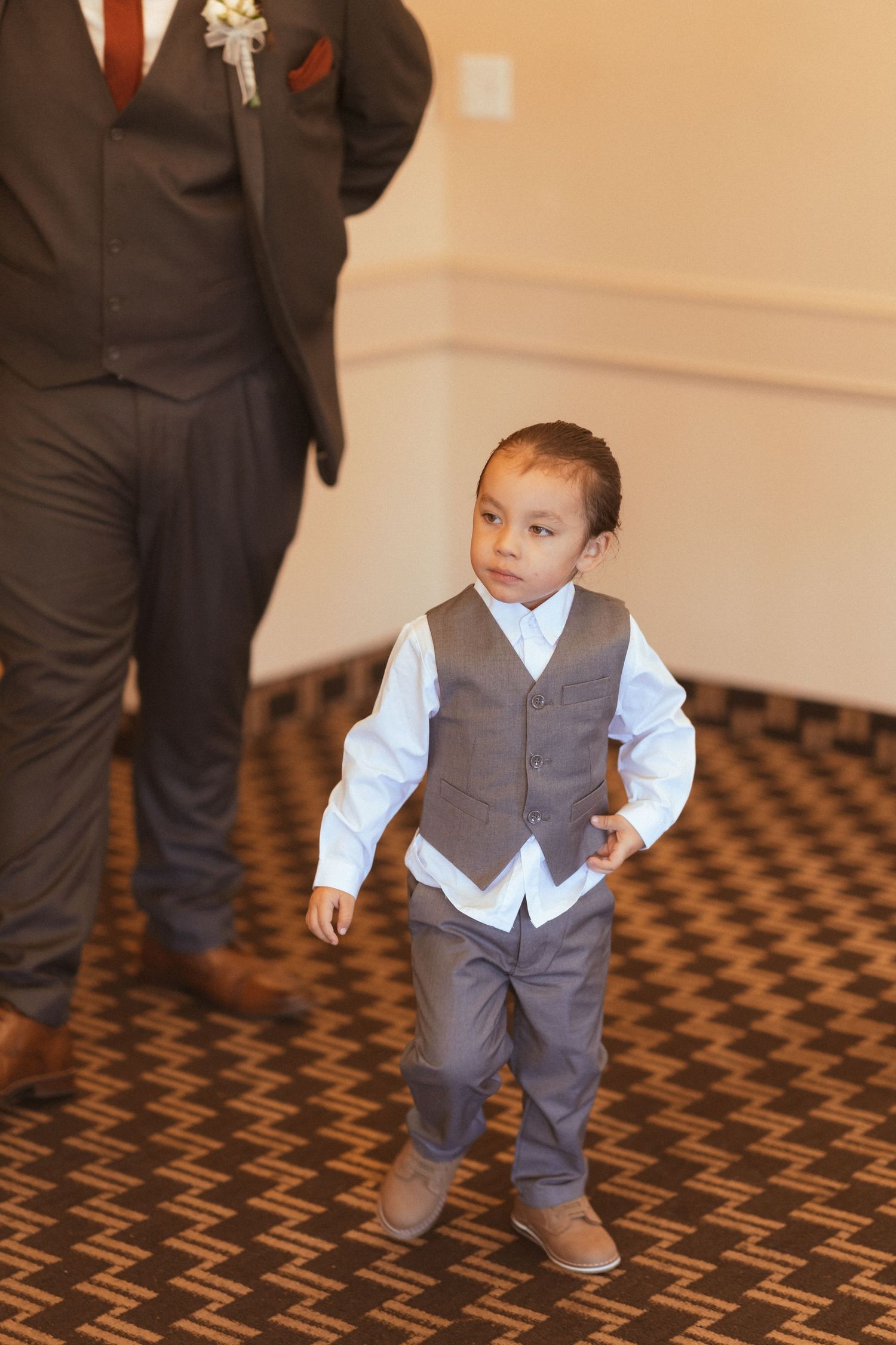 A young child in formal attire stands on a patterned carpet wearing a vest and dress shirt.