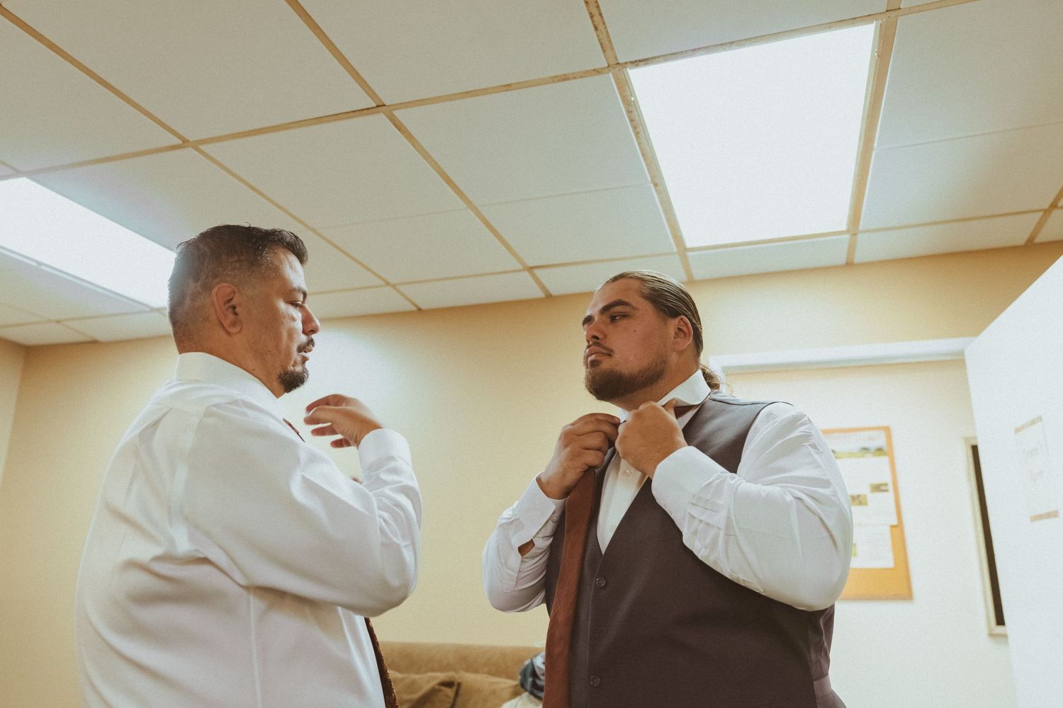Two people in formal attire help each other adjust their ties before an event.