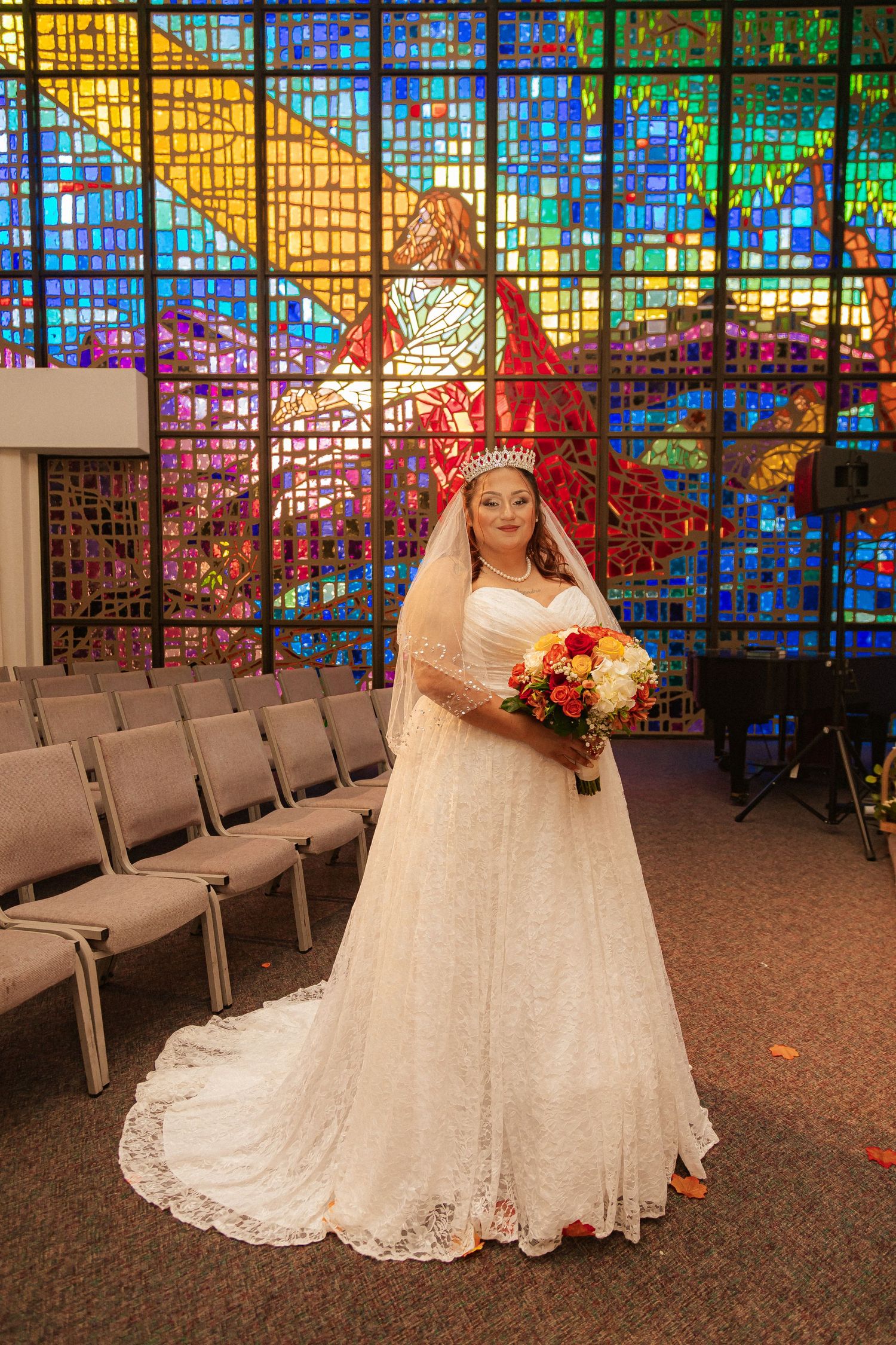Bride in lace wedding gown poses with bouquet against backdrop of colorful stained glass church windows.