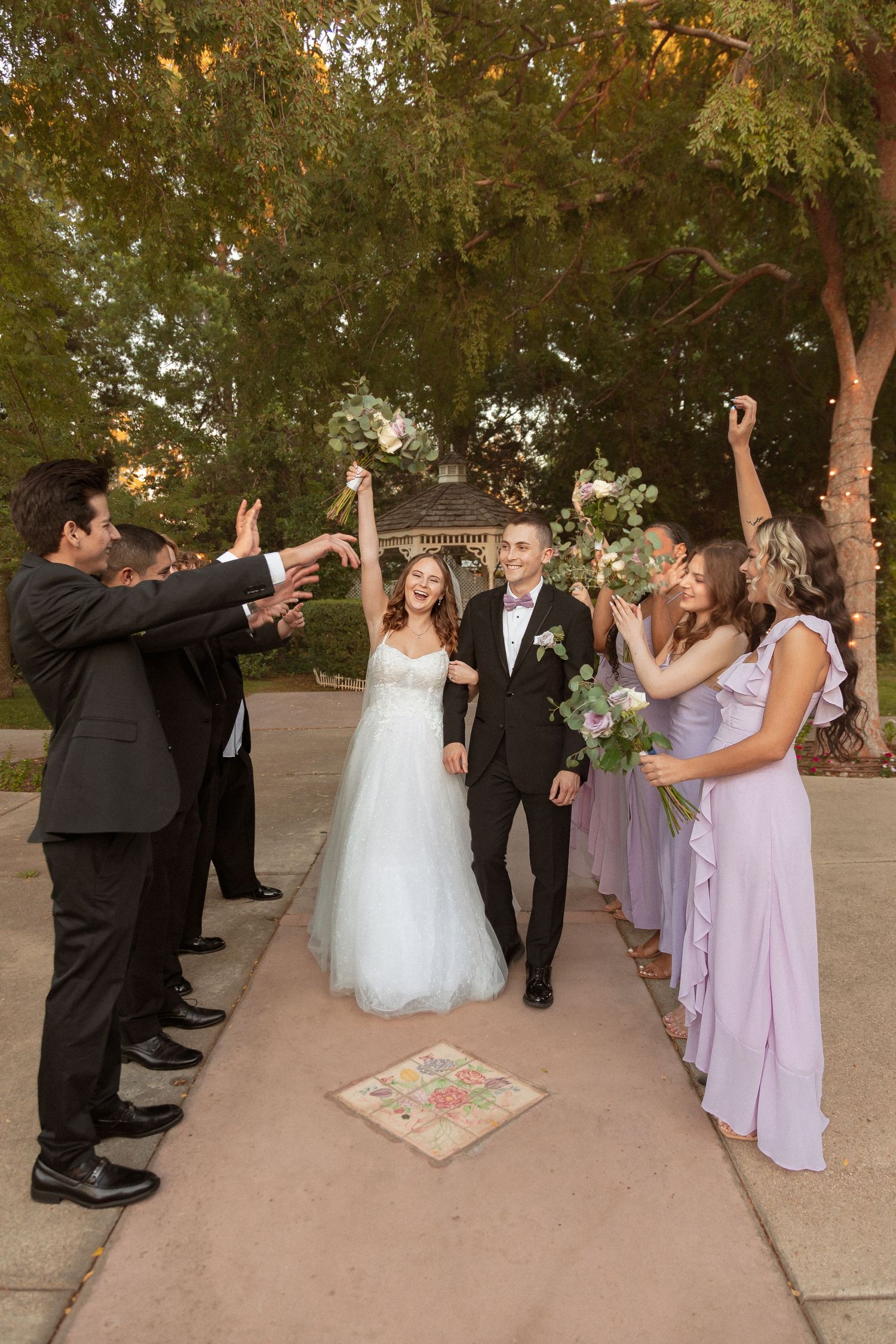 Wedding party celebrates with raised hands on outdoor pathway during sunset ceremony.