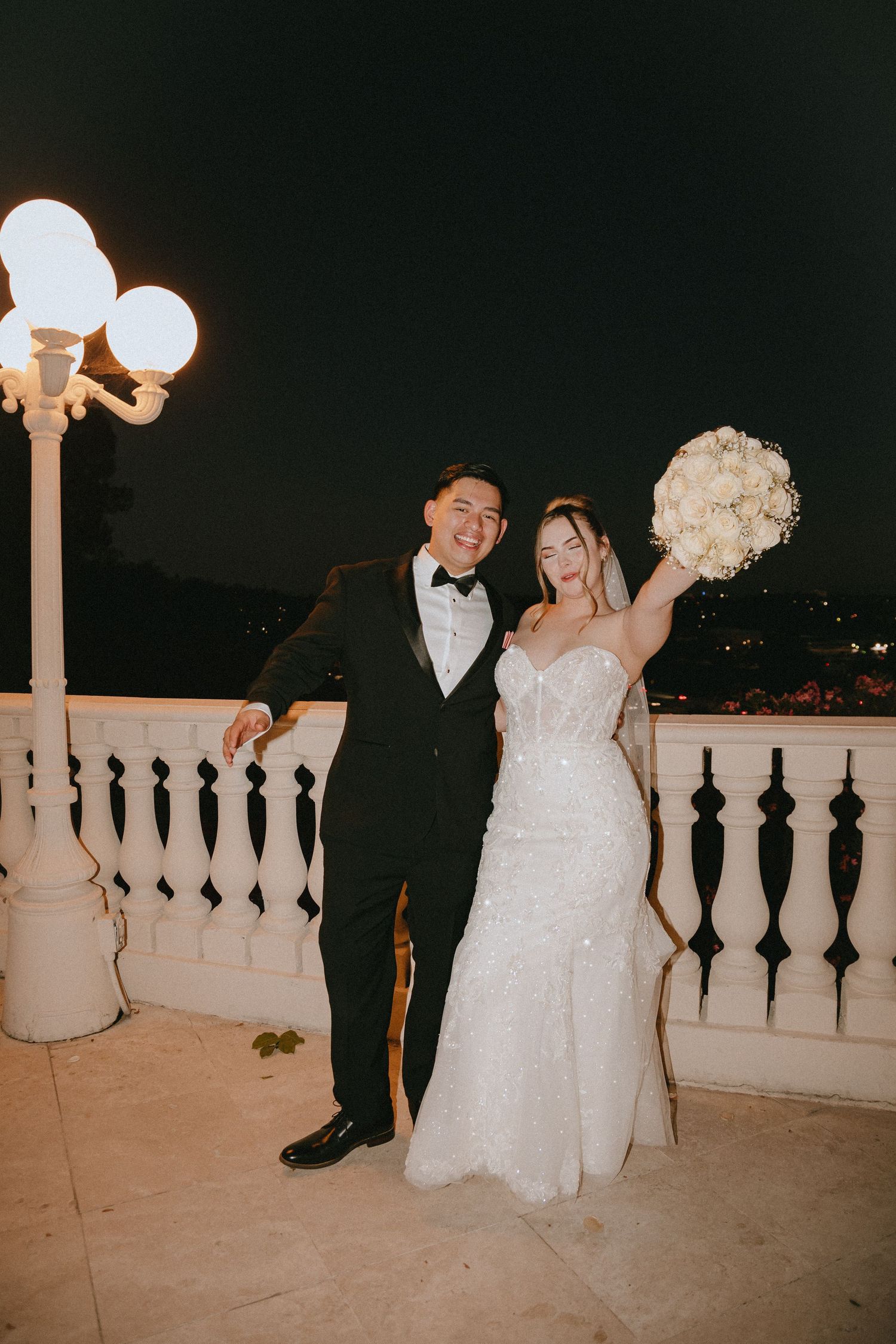 Newlyweds share a joyful moment on an illuminated terrace with a white floral bouquet.