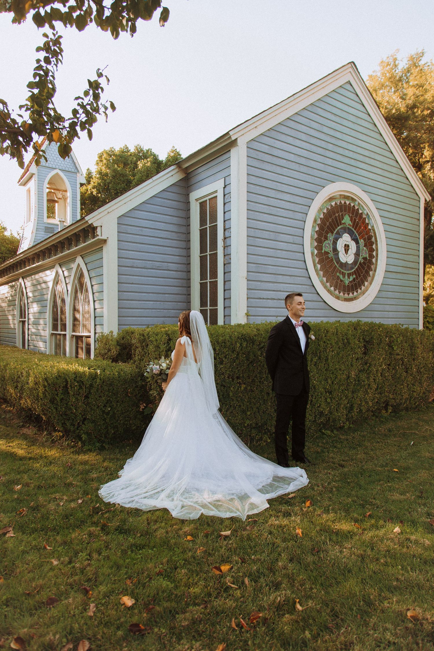 Wedding couple stands separately outside blue chapel with distinctive round window and hedges.