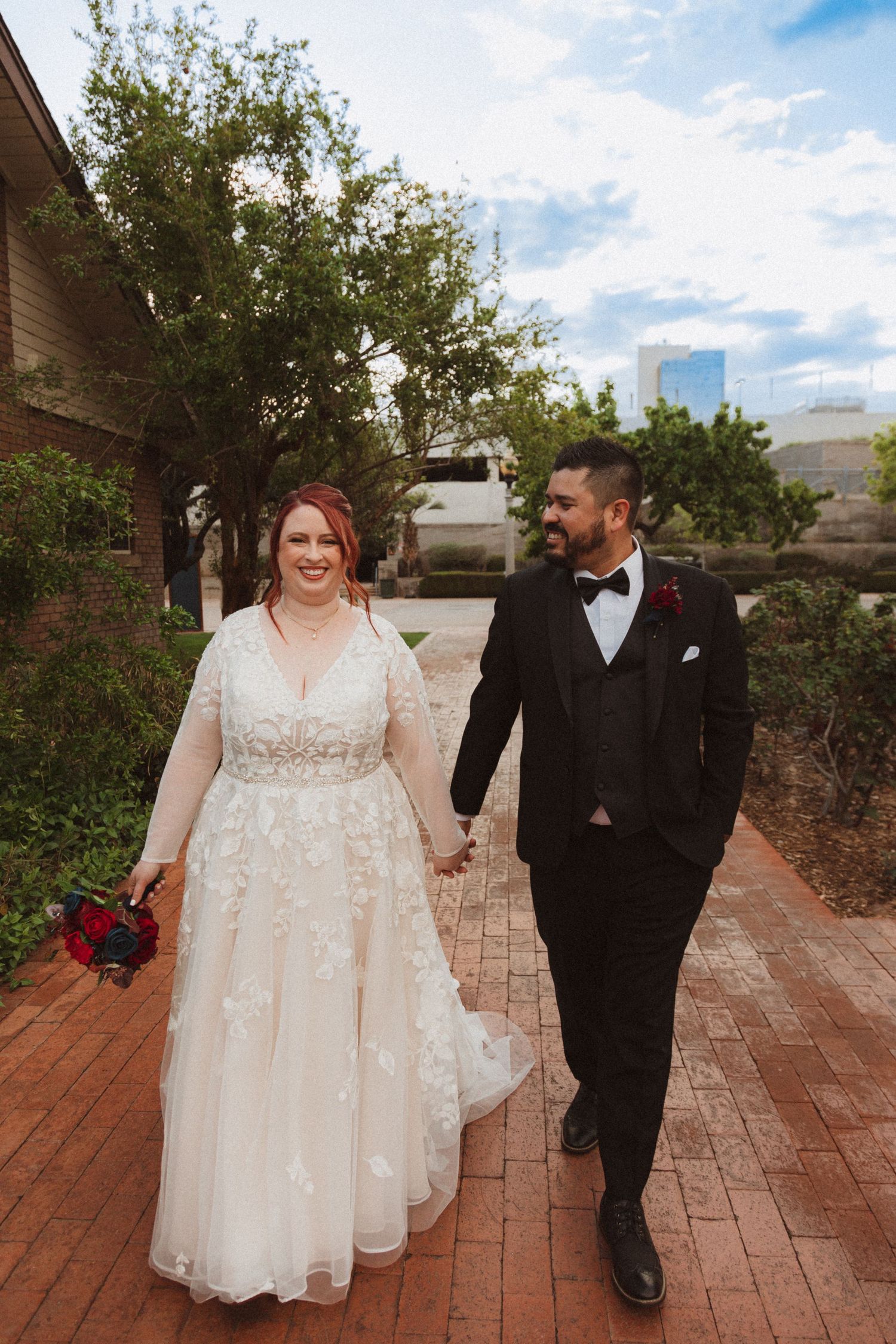 Newlyweds share a joyful moment while walking on a brick path with urban buildings in background.