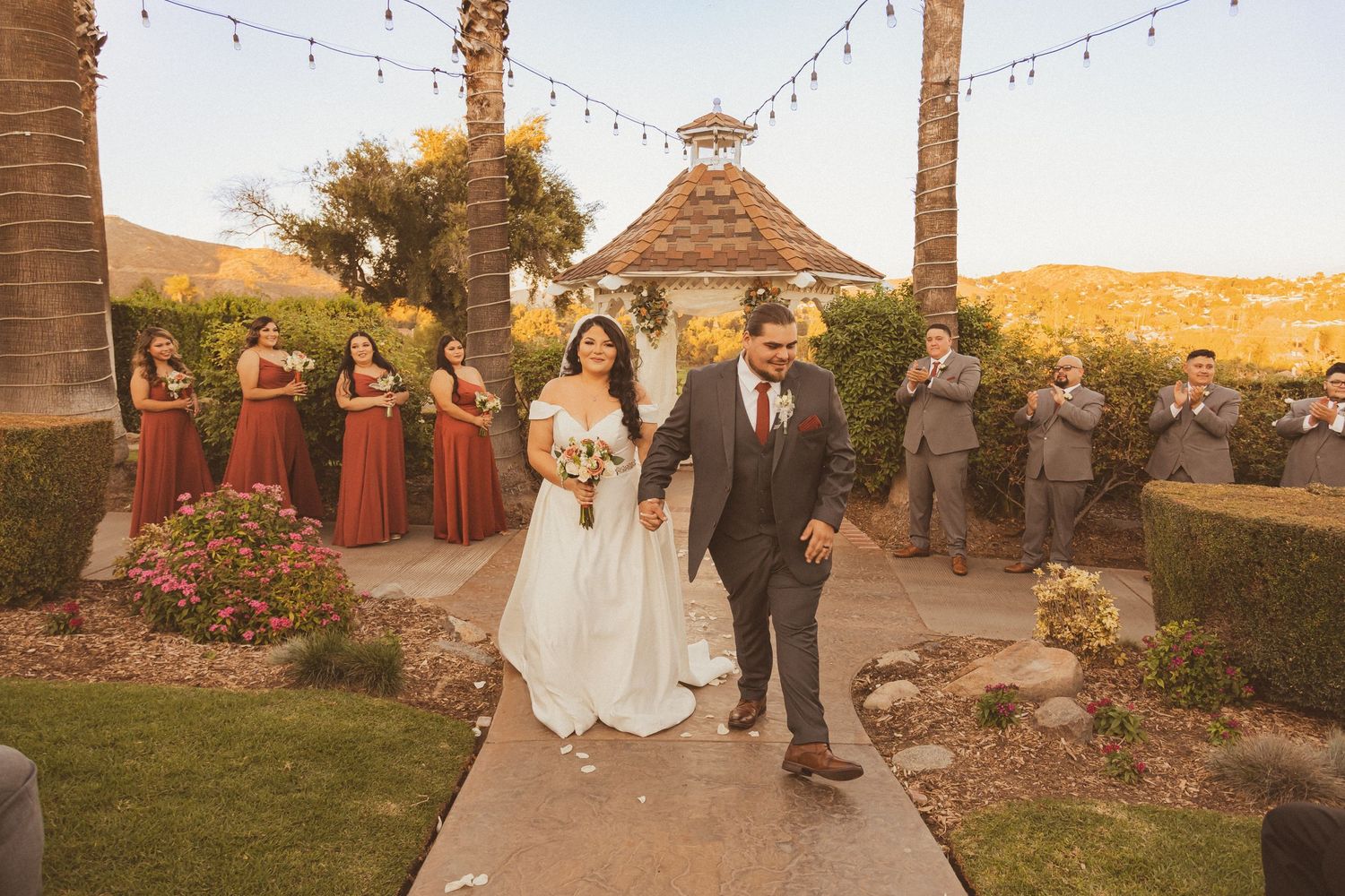 Wedding party exits ceremony under string lights with rustic gazebo and mountains behind.
