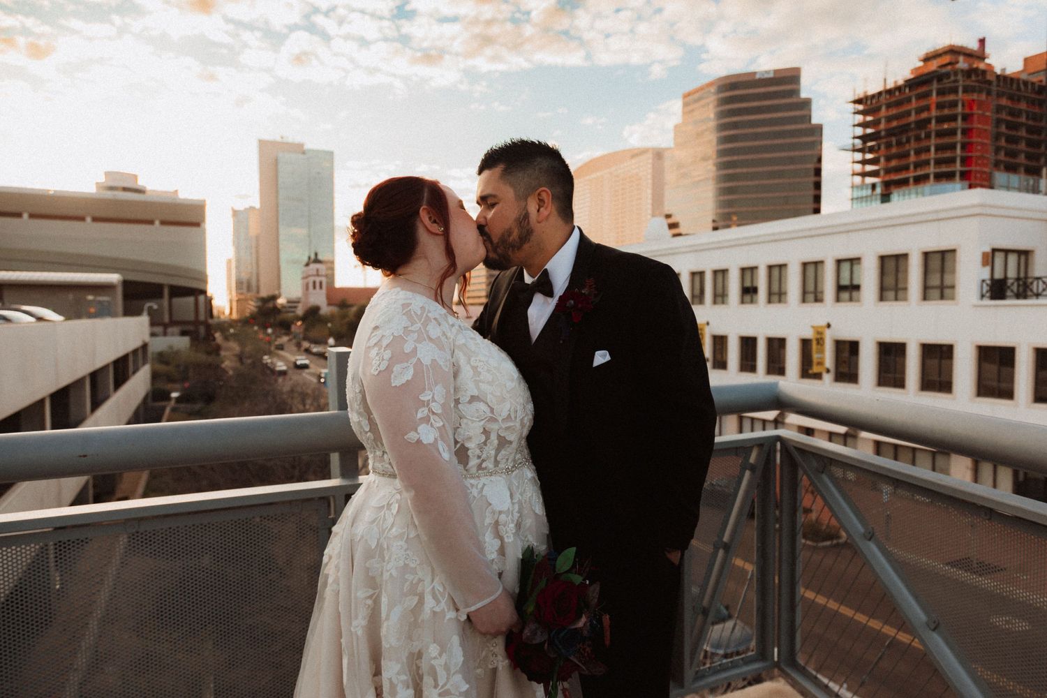 A romantic wedding couple embraces on a downtown rooftop at sunset with city buildings in the background.