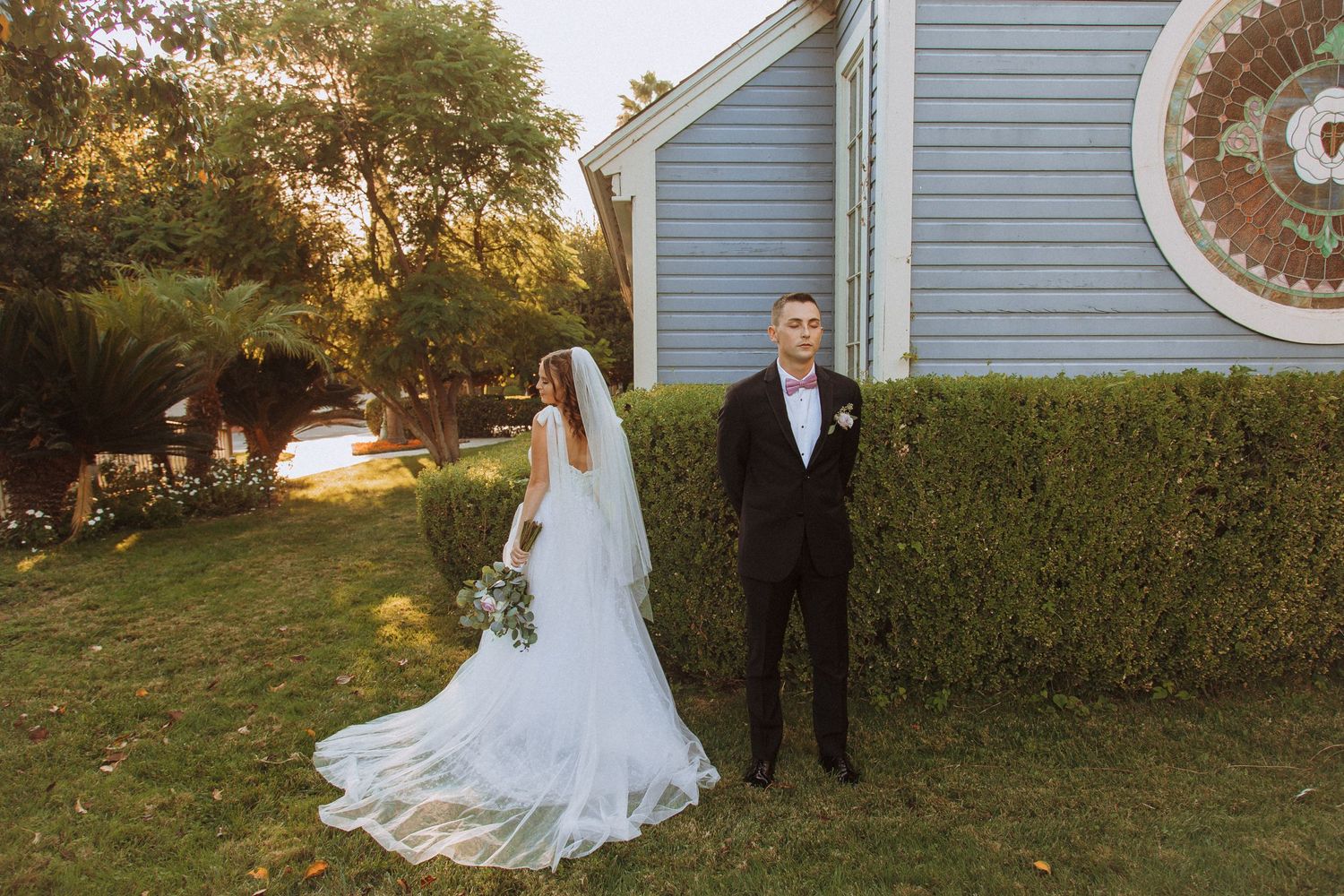 Wedding couple stands by blue chapel during golden hour with bride holding floral bouquet.