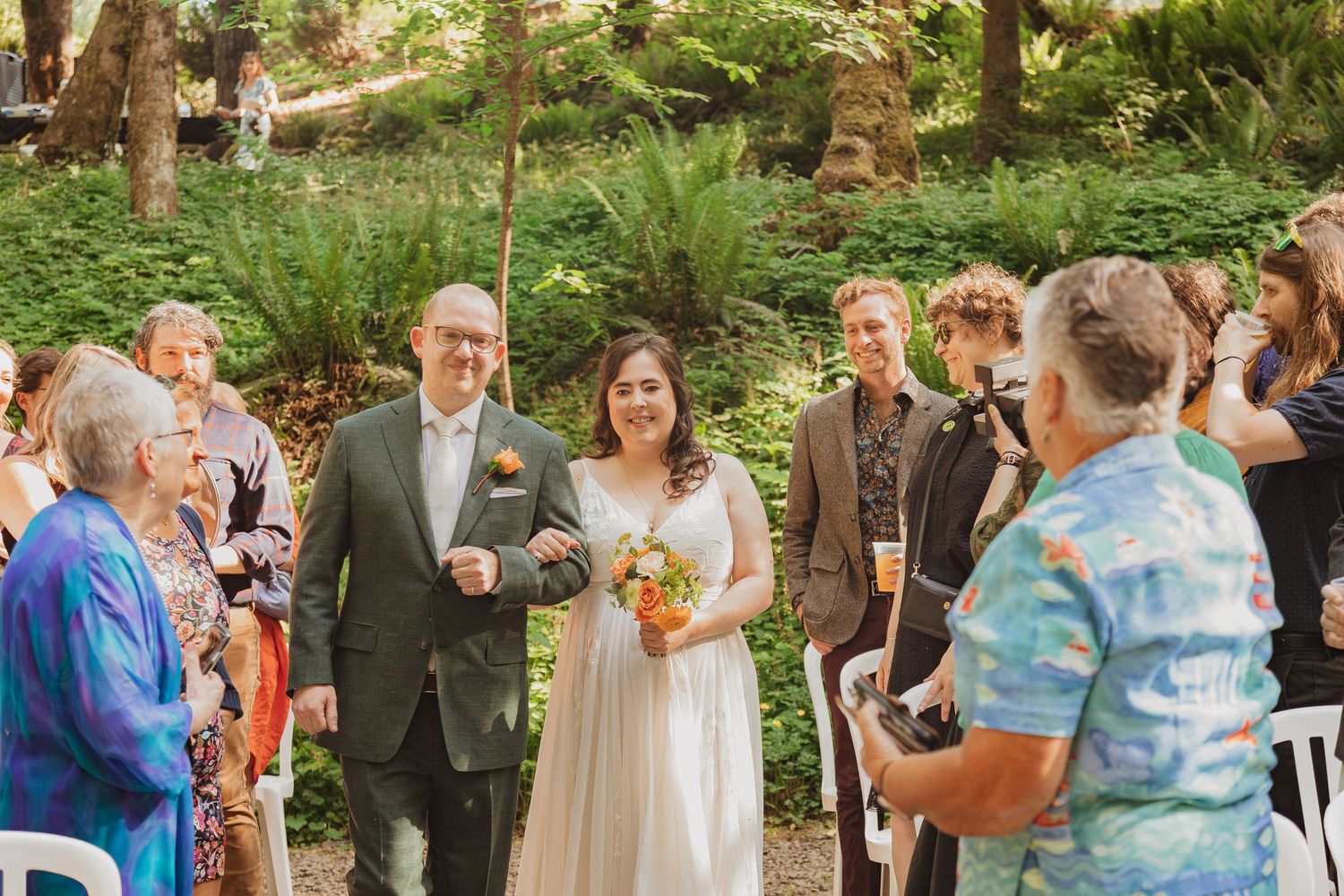 An outdoor wedding ceremony with guests gathered in a sunlit forest clearing.