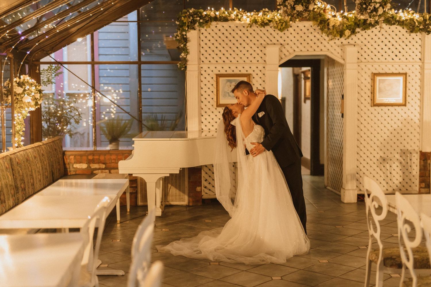 Couple shares tender moment under white lattice archway with fairy lights and greenery in warm lighting.