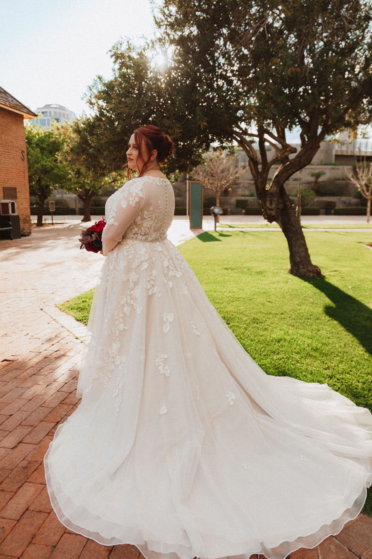 A flowing white wedding gown with intricate lace details shown from behind on brick pathway.