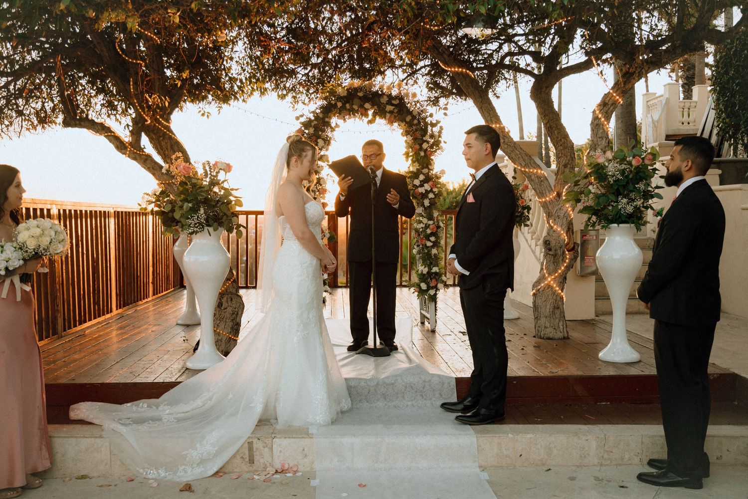 A wedding ceremony takes place under trees decorated with fairy lights and white floral arrangements.