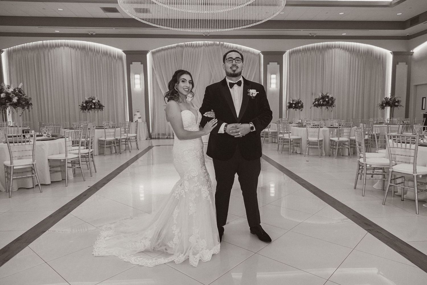 Black and white photo of couple standing in grand ballroom with elegant table settings.