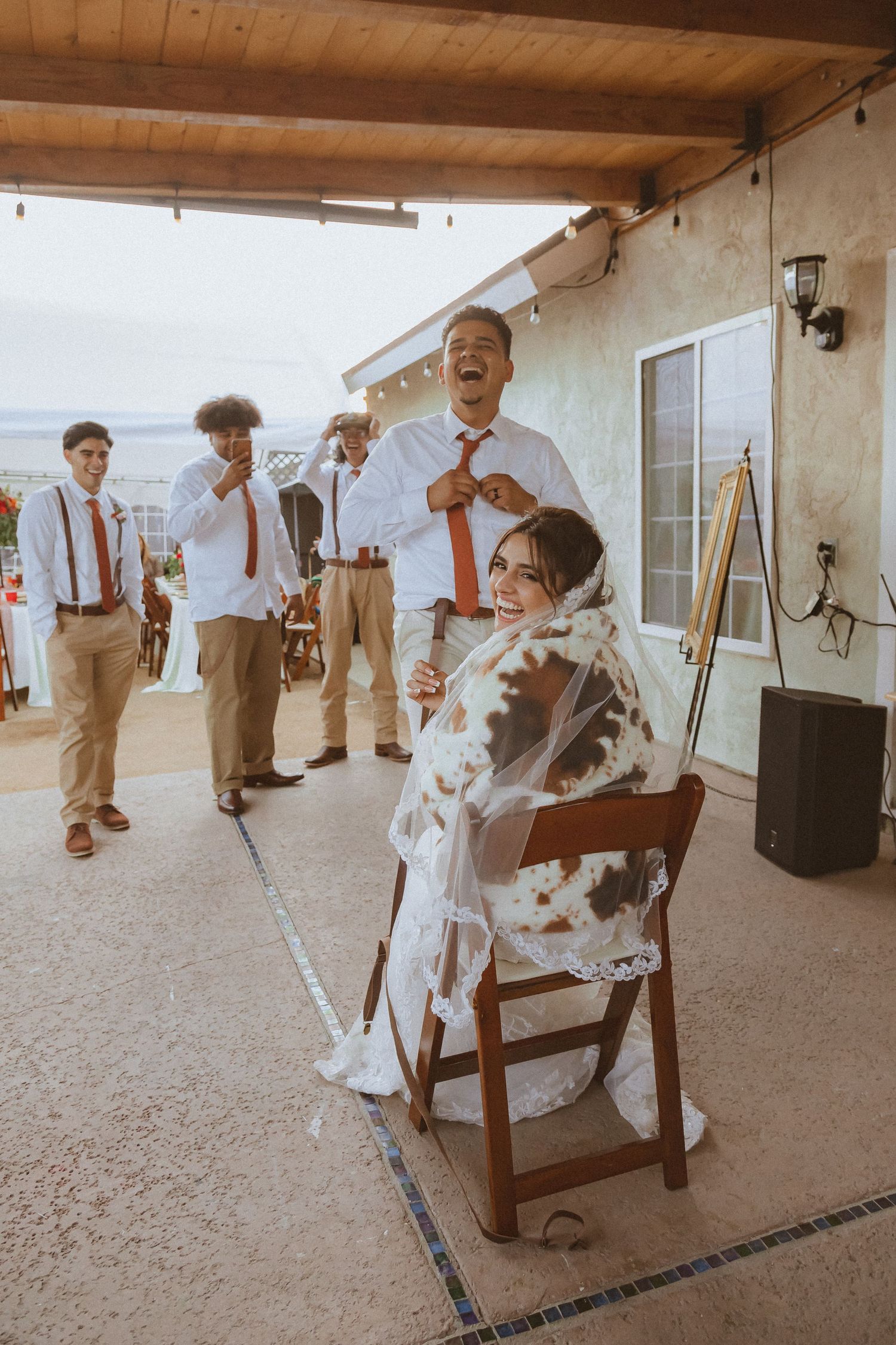 Wedding party members in suspenders and ties laugh during a reception celebration outdoors.