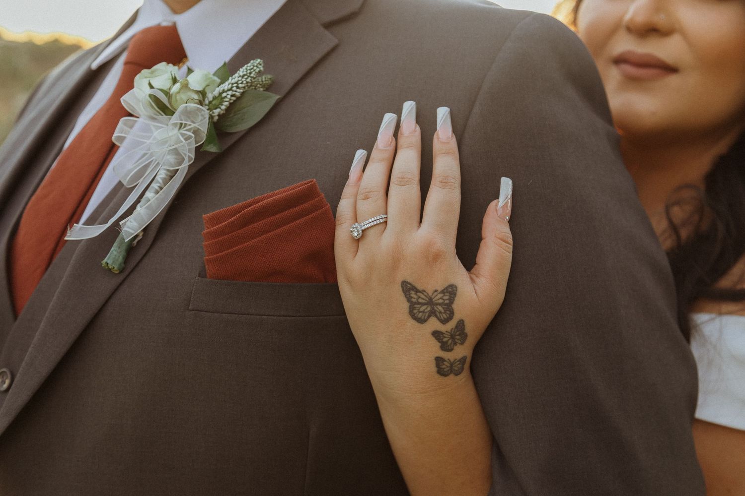 Hand with butterfly tattoo and engagement ring resting on brown suit jacket with floral boutonniere.