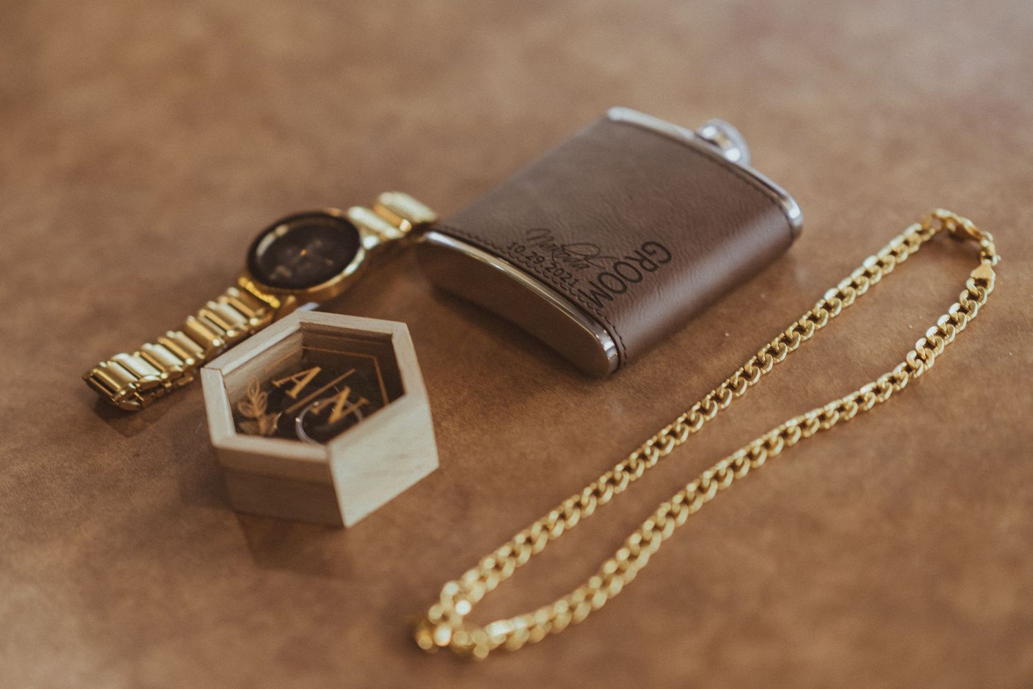 Brown leather flask, gold watch, hexagonal container and gold chain necklace arranged on brown leather surface.