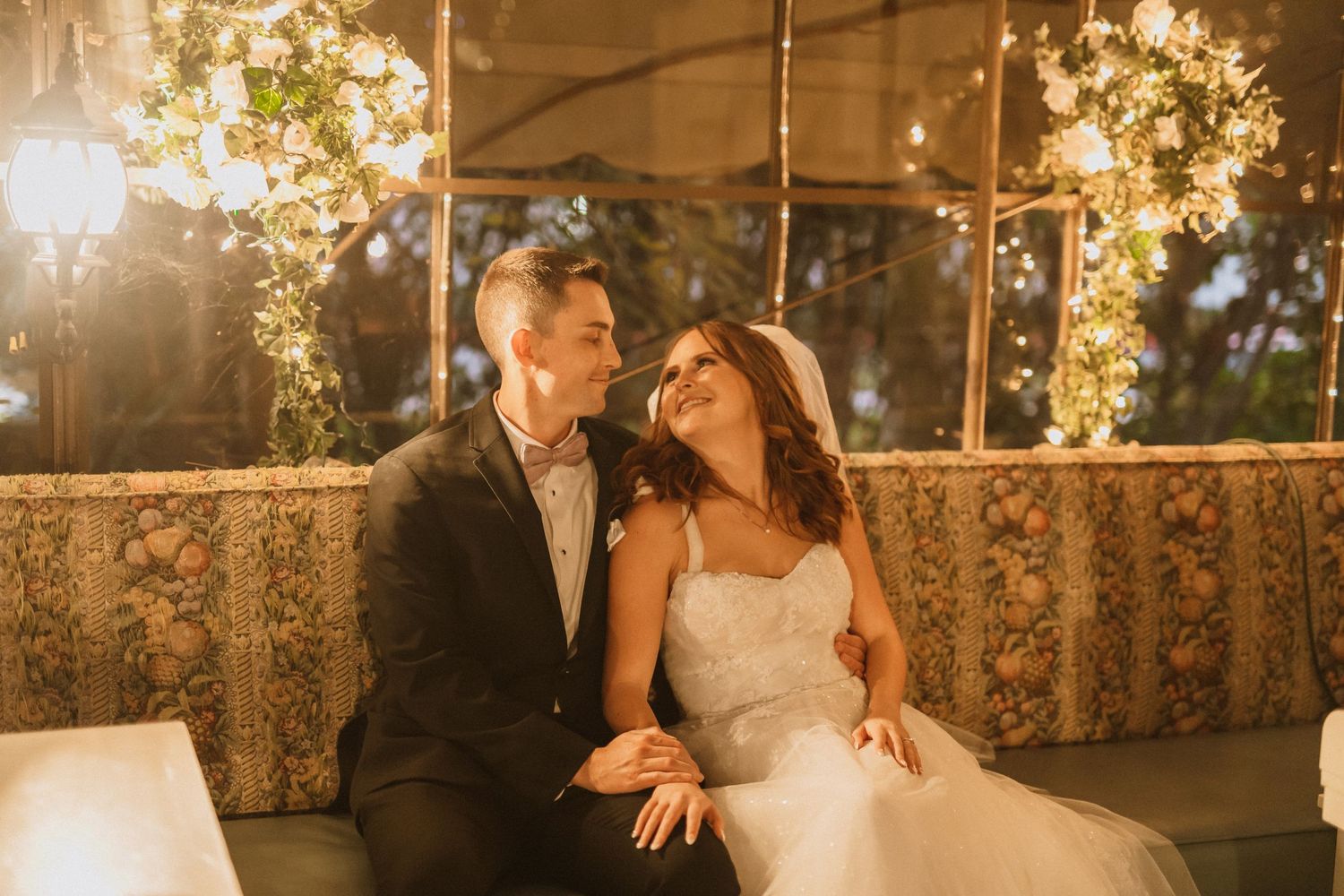 Sunset wedding portrait showing joyful couple beneath warm glowing lights and floral decorations.