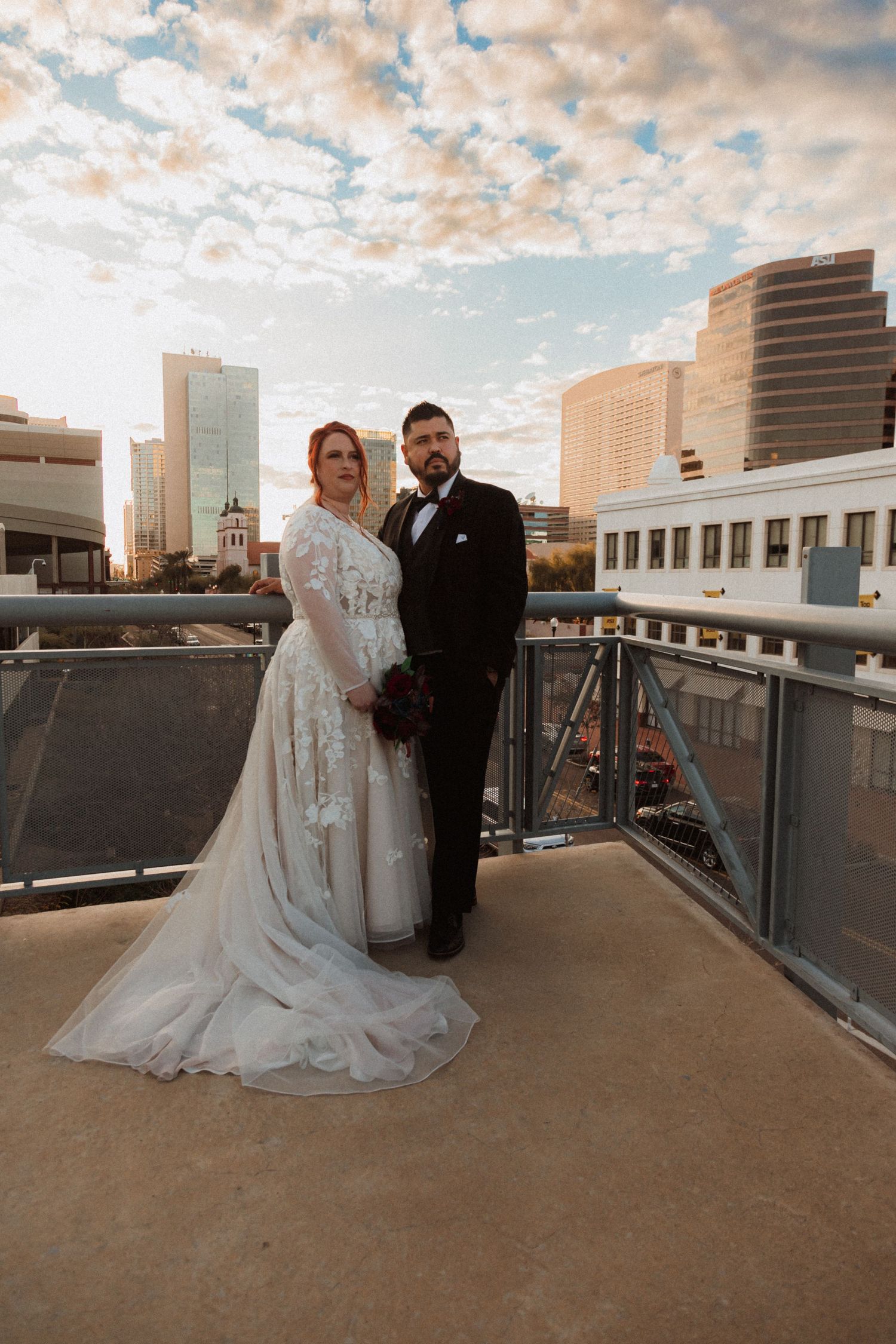 Bride in lace gown and groom in tux share intimate moment on city rooftop at dusk.