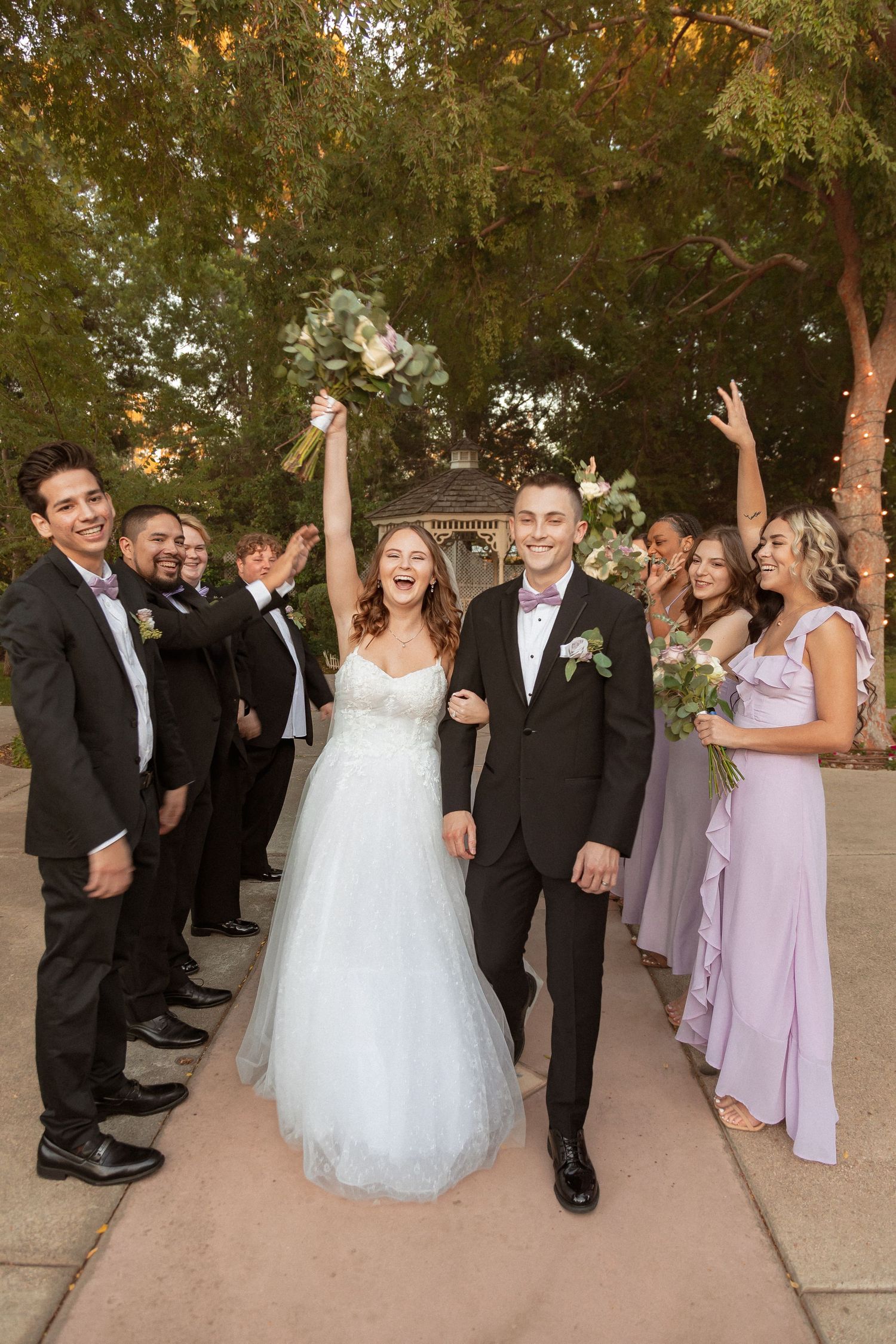 Joyful wedding party celebrates with raised arms on outdoor pathway.