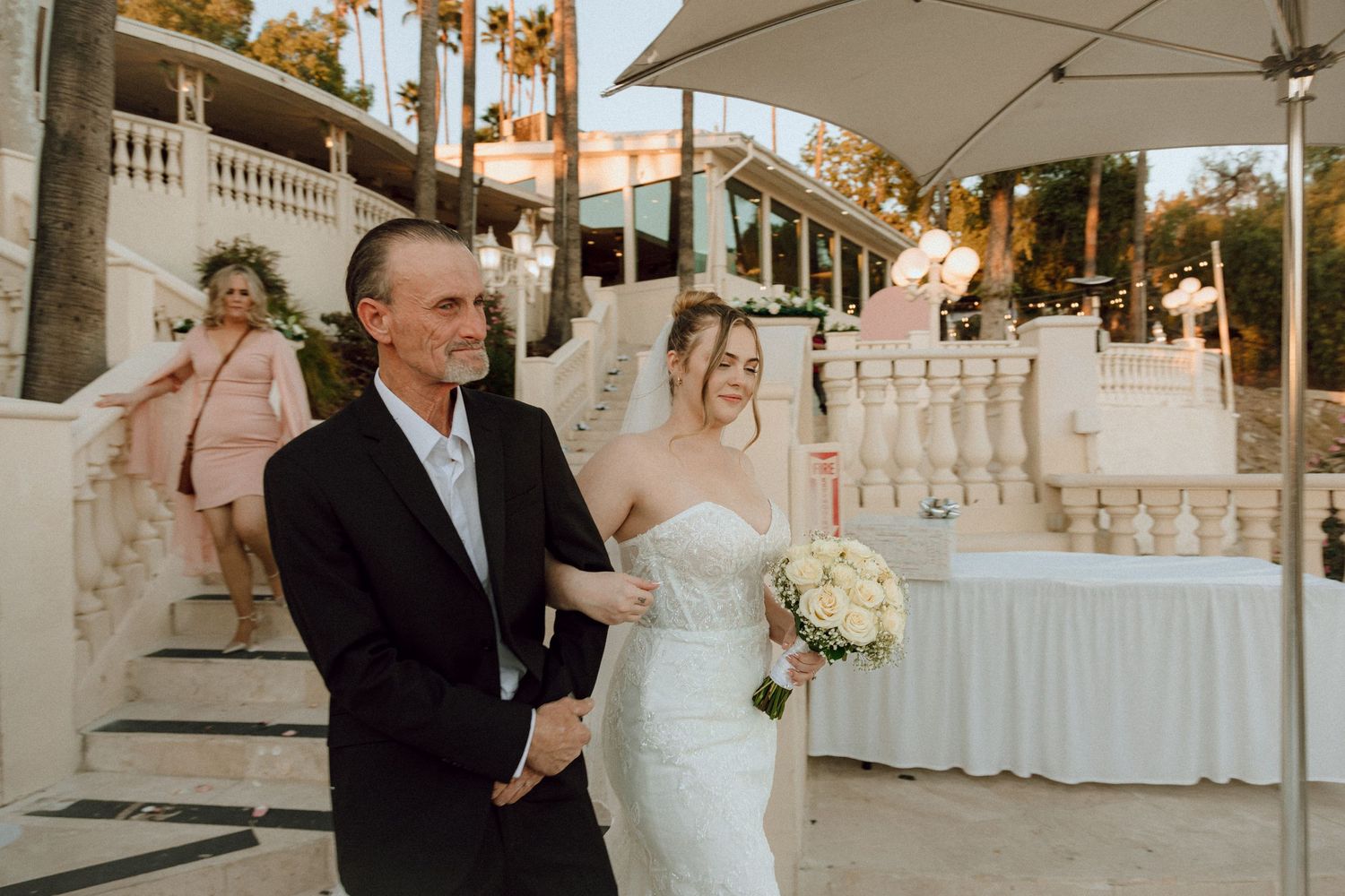 Wedding party walking down white stone steps with tropical landscaping and reception tables visible.