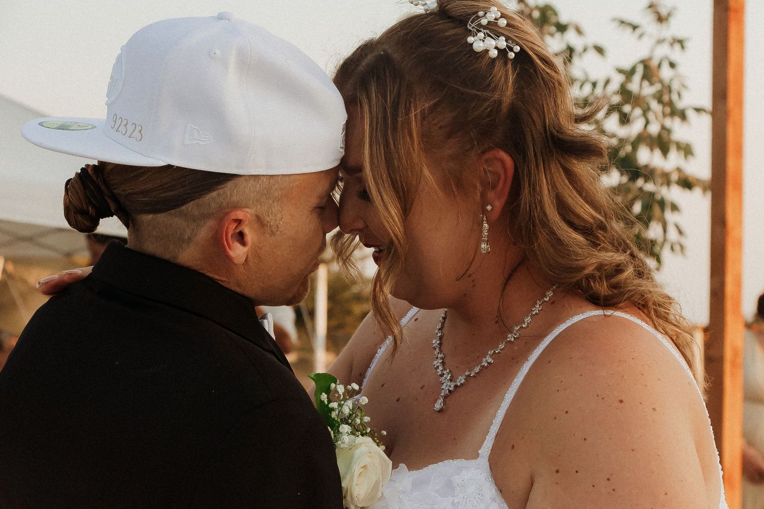 A tender wedding moment captured outdoors with the groom wearing a white baseball cap.