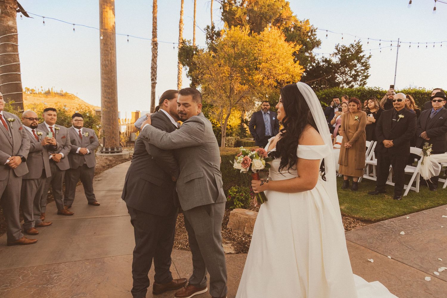 Wedding party celebrates outdoors under string lights and palm trees during golden hour.