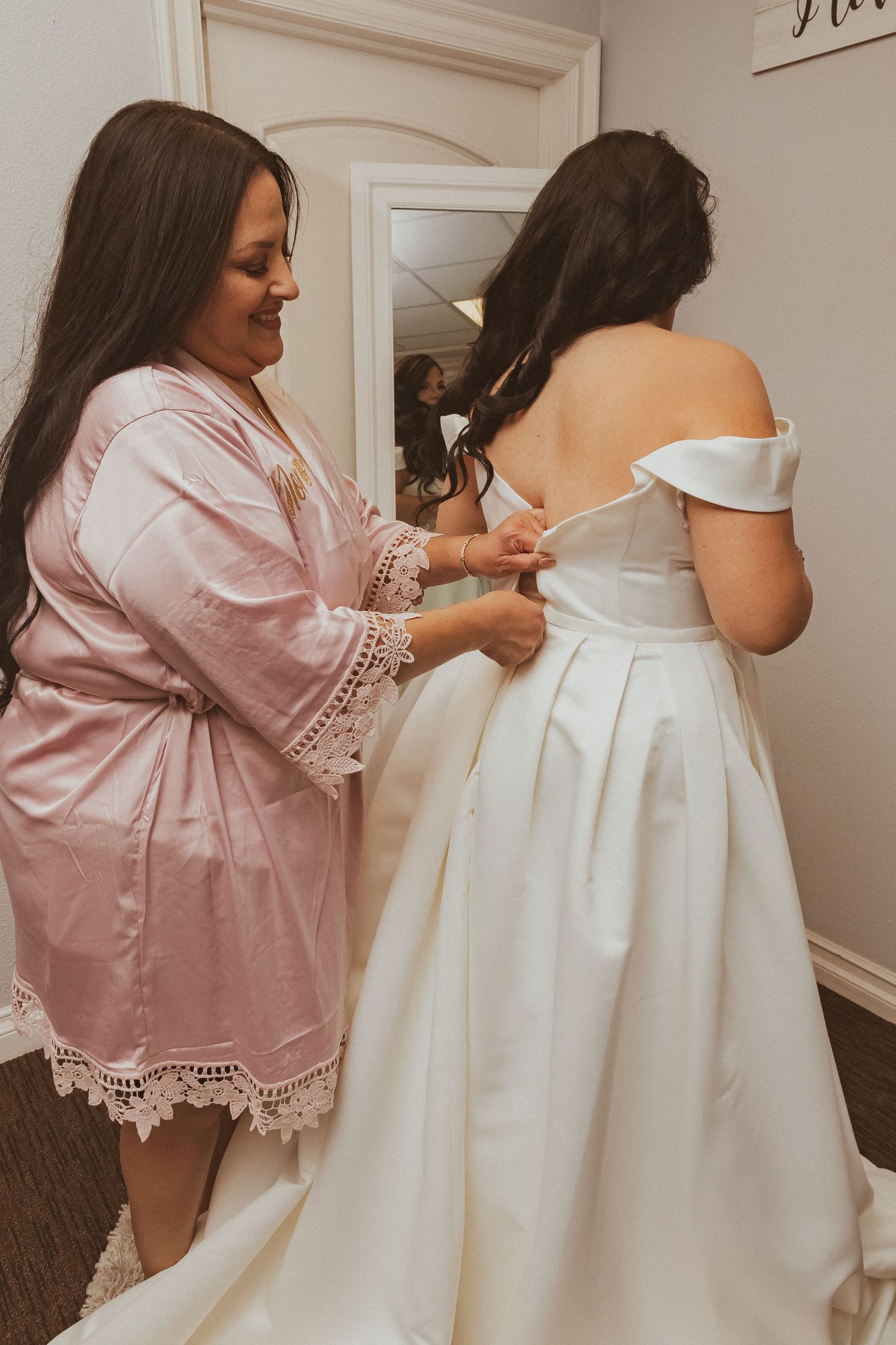 Woman in pink lace robe helps adjust a classic white off-shoulder wedding dress during final fitting preparations.