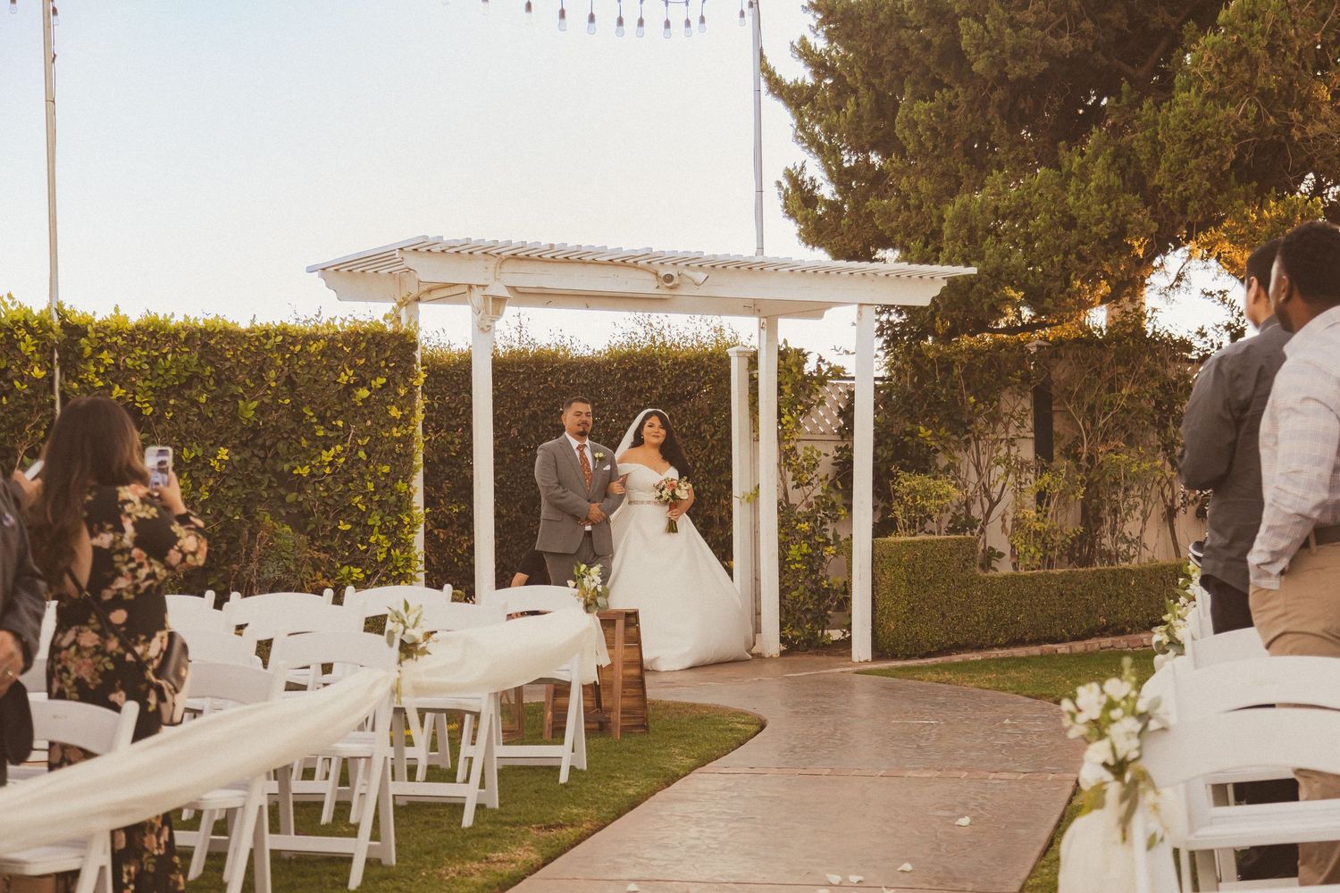 Romantic outdoor wedding ceremony under a white pergola with string lights and garden backdrop.