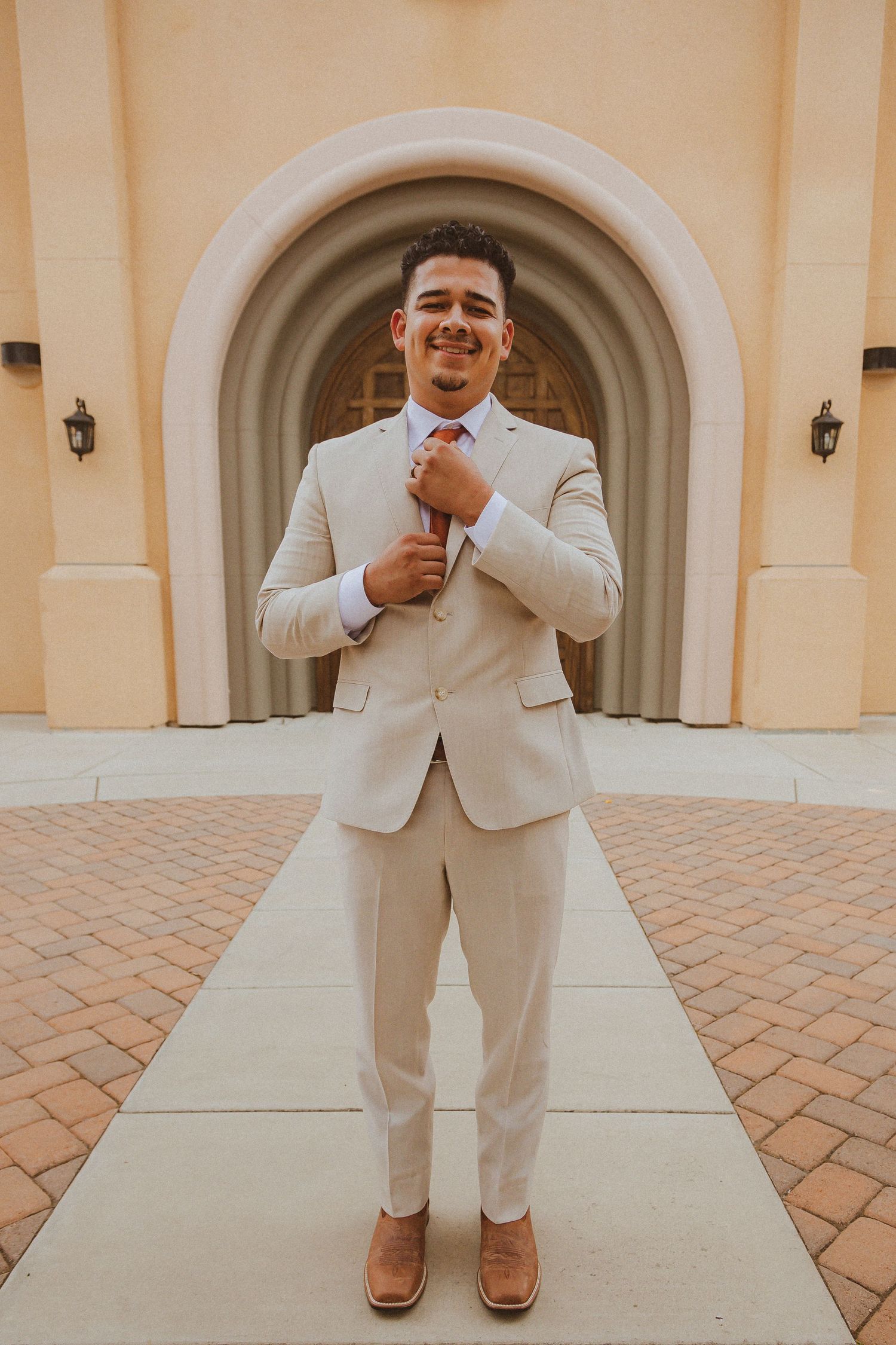 Full-length portrait of wedding attire featuring beige suit and orange tie against arched entrance.